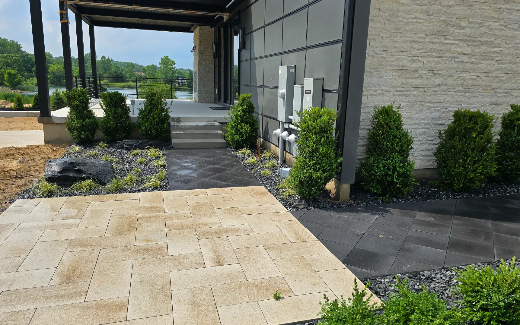 Modern outdoor walkway with light and dark stone tiles, landscaping with green shrubs, and a view of a lake and trees in the background.