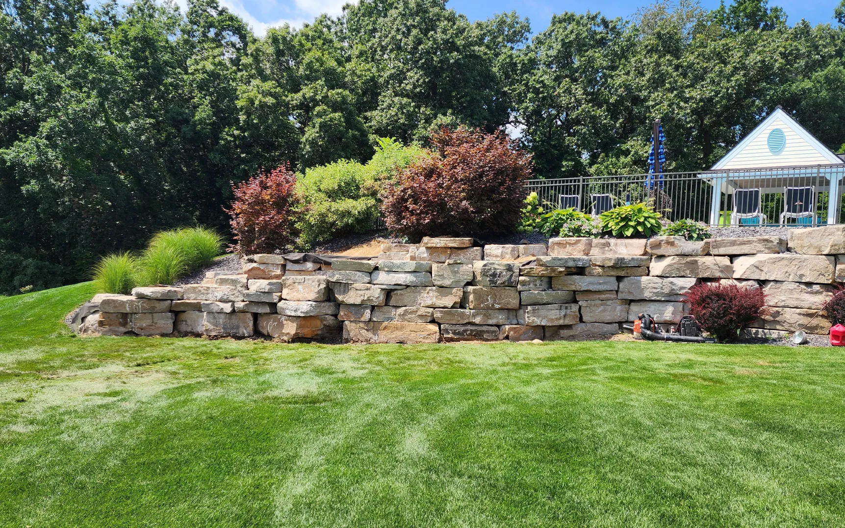 Well-maintained backyard with green lawn, tiered stone retaining wall, shrubs, and a small white pavilion with chairs behind a black metal fence.