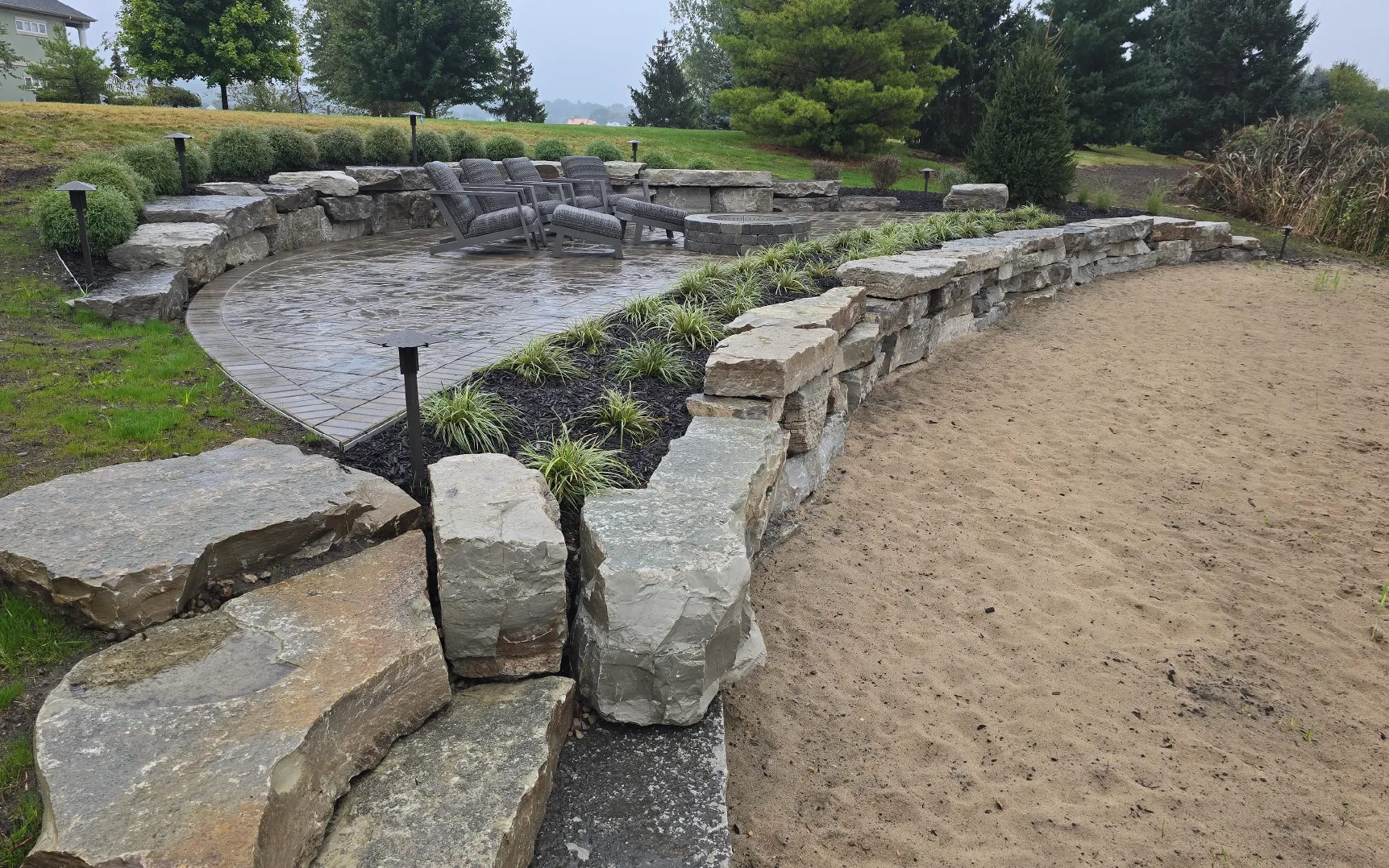 Outdoor patio area with stone retaining walls, wicker chairs around a fire pit, and sand-covered ground adjacent to the patio.