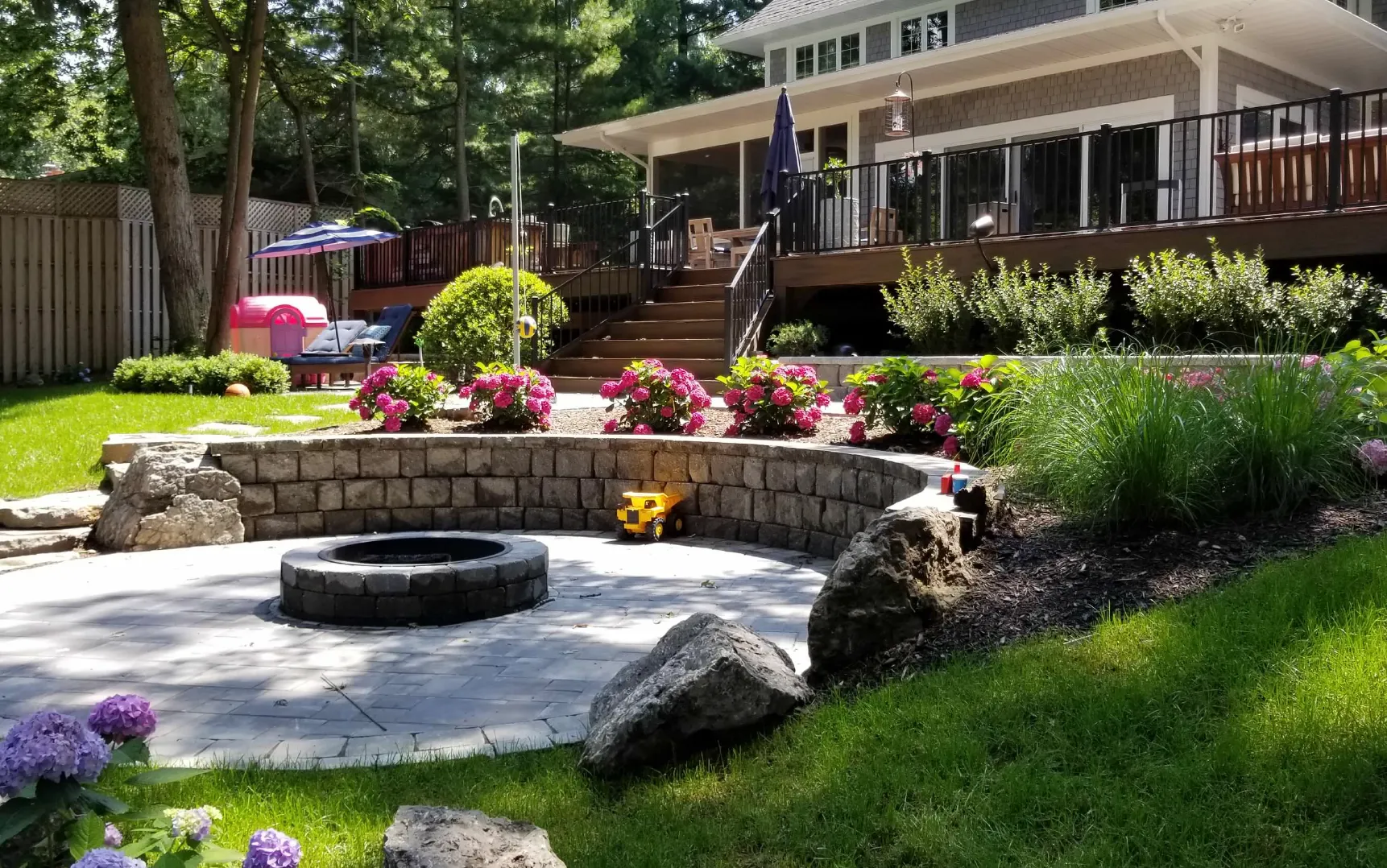 Backyard with a stone fire pit, curved retaining wall with pink flowers, green grass, and a wooden deck with stairs leading up to a house.