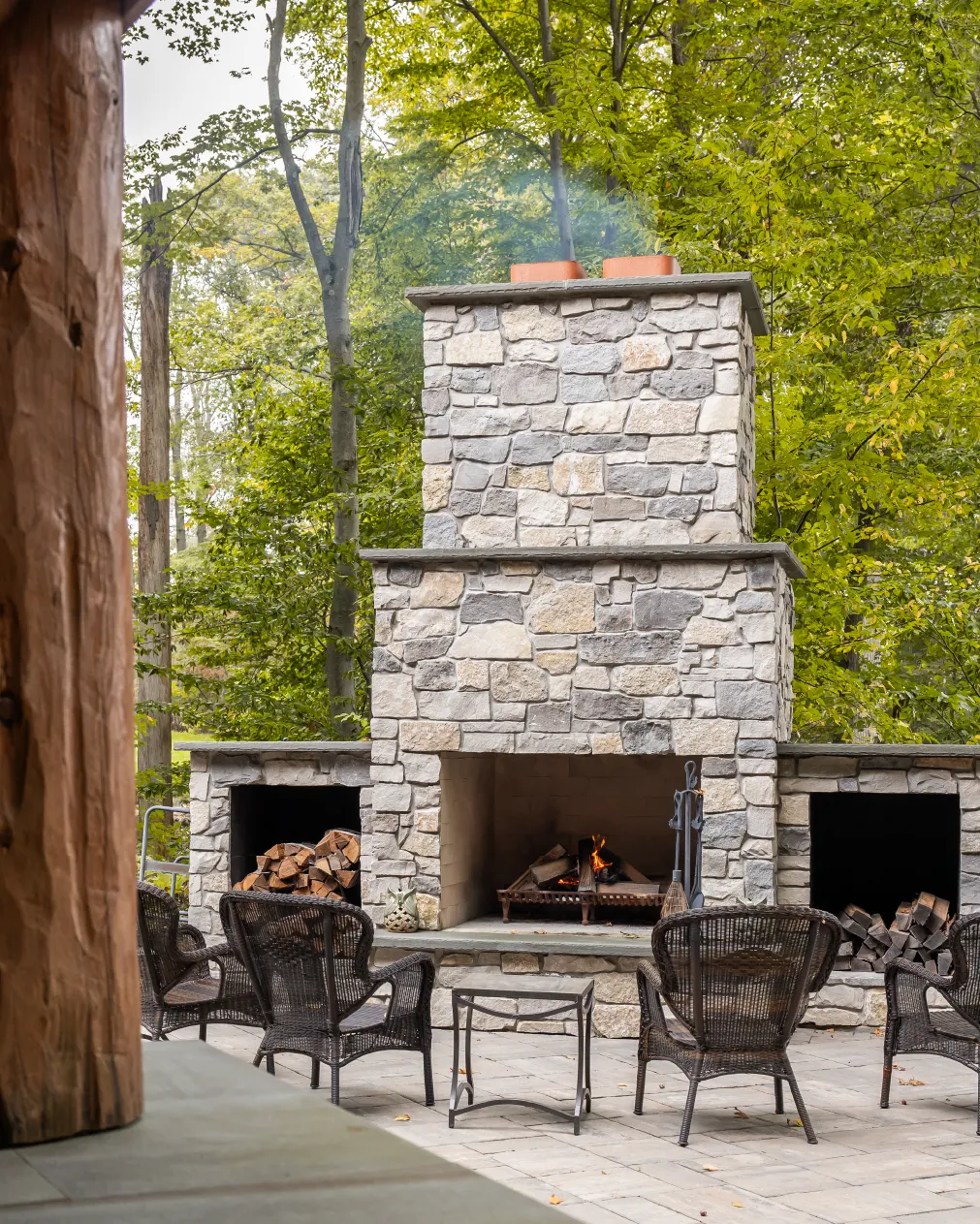 Outdoor stone fireplace with fire burning, surrounded by wicker chairs and firewood storage, set against green trees.
