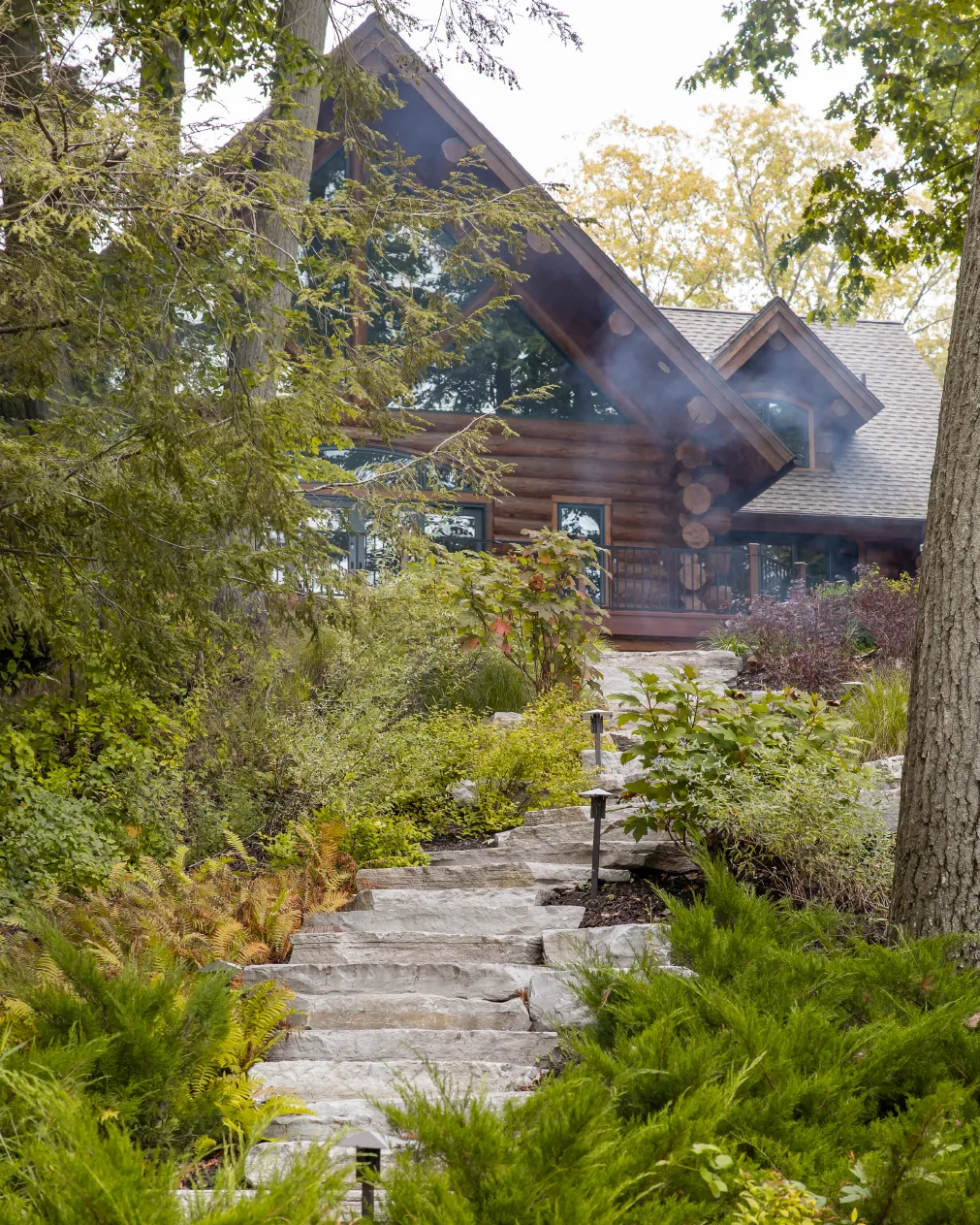 Stone steps leading through lush greenery to a rustic log cabin with large windows and a gabled roof.