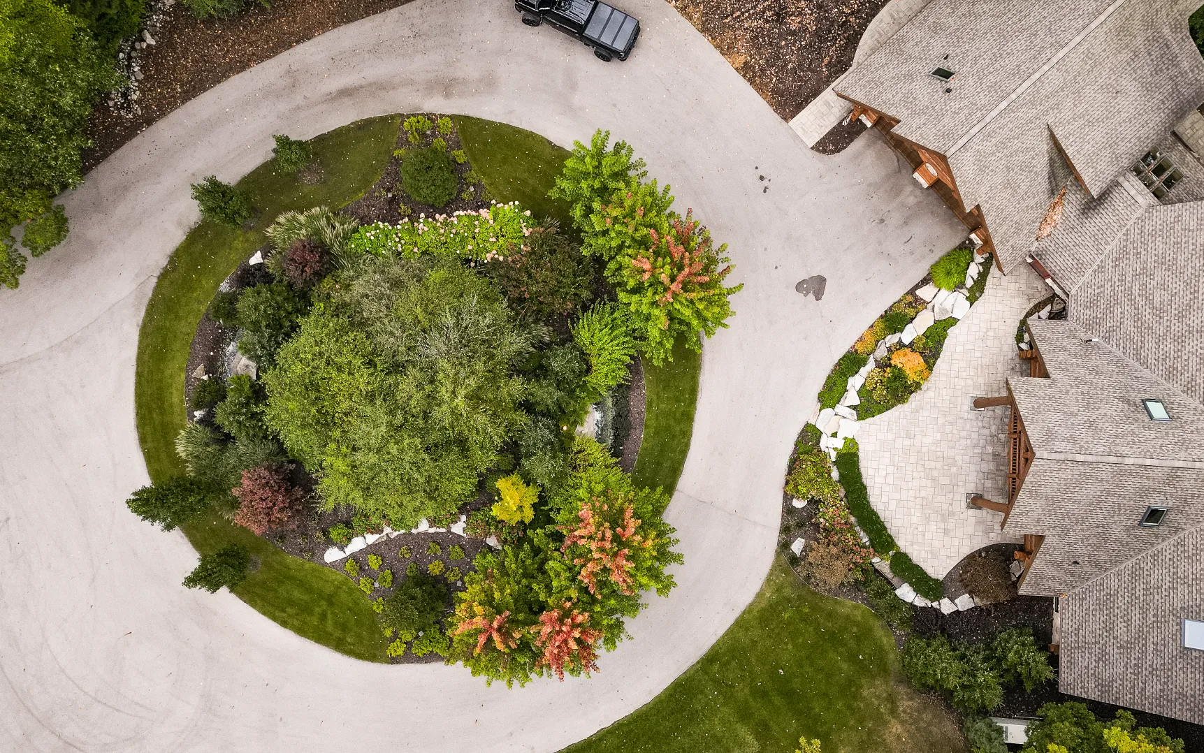 Aerial view of a circular driveway surrounding a landscaped garden with various trees and shrubs next to a house with a gray roof.