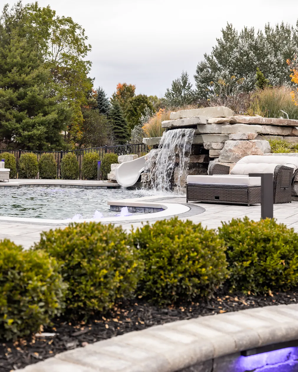 Swimming pool with a slide and waterfall feature next to cushioned outdoor lounge chairs surrounded by shrubs and trees.