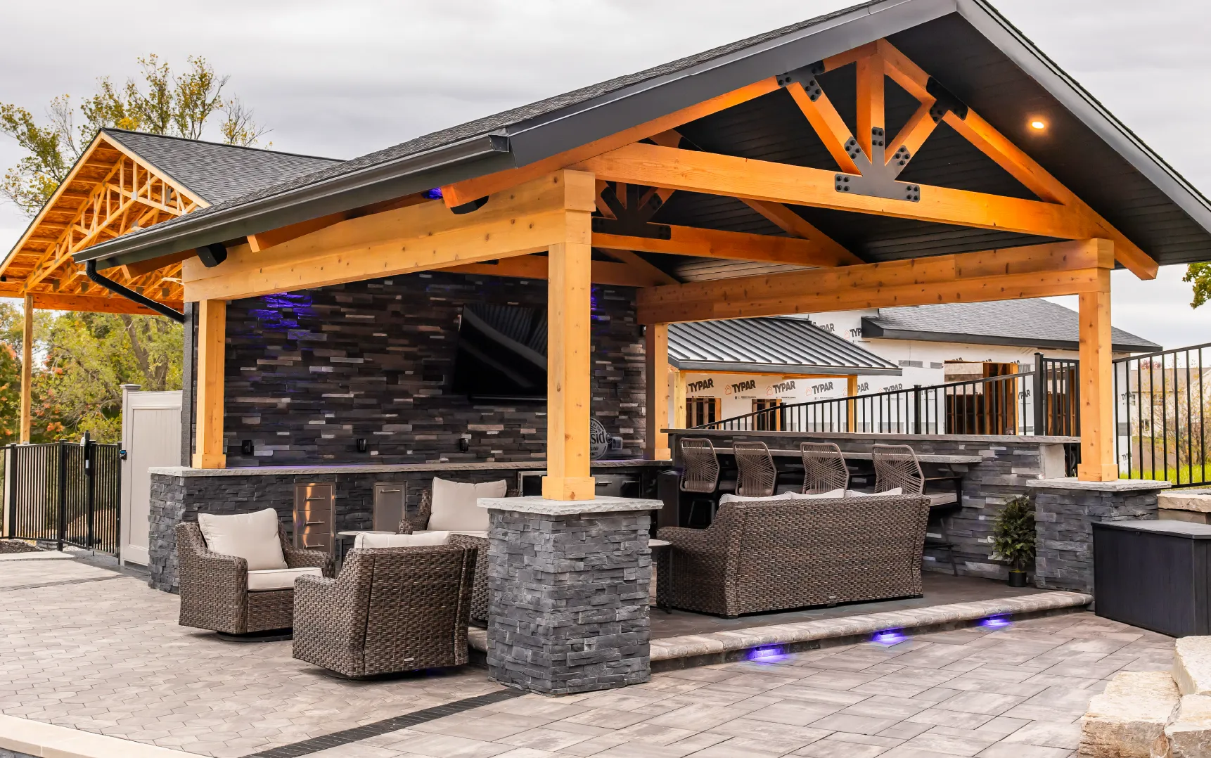 Outdoor covered patio with wooden beams, stone pillars, wicker seating, bar stools, and a mounted television.