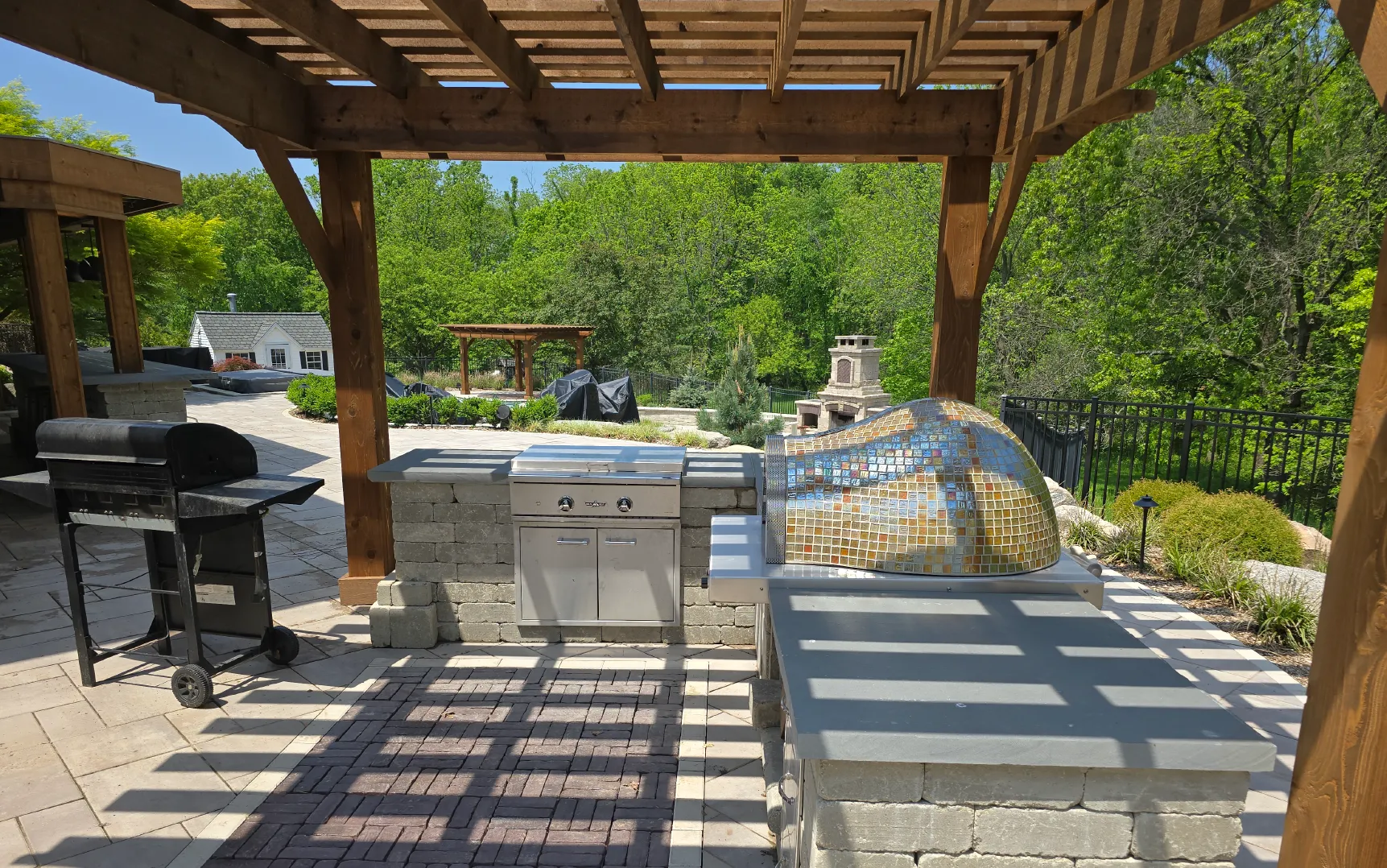 Outdoor kitchen under a wooden pergola with a charcoal grill, built-in stainless steel gas grill, and a mosaic-tiled pizza oven.