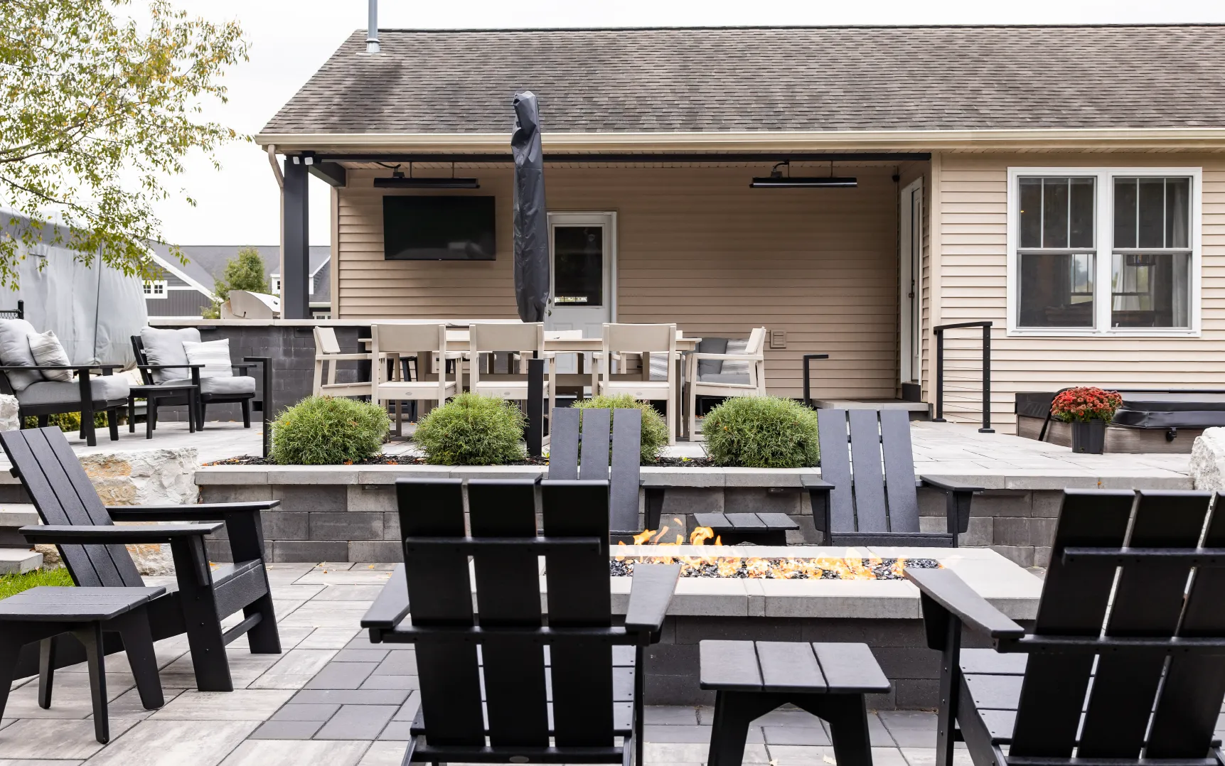 Outdoor patio area with black Adirondack chairs around a rectangular fire pit, beige dining table with chairs, and seating under a covered porch.