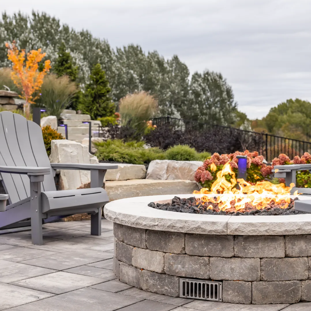 Outdoor stone fire pit with flames surrounded by gray Adirondack chairs on a tiled patio with landscaped greenery in the background.