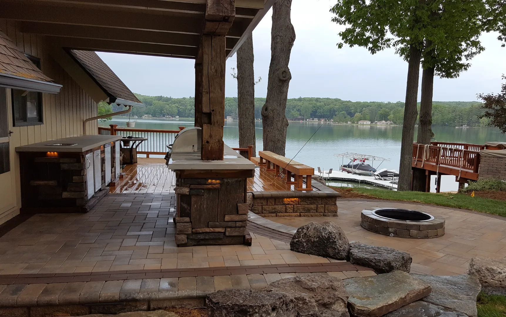 Outdoor patio with built-in grill, stone fire pit, wooden bench, overlooking a lake with docks and boats.