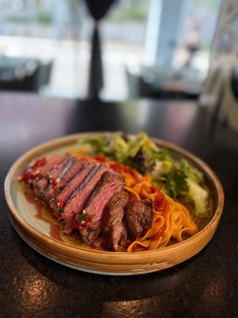 Plate of sliced medium-rare steak served over tagliatelle pasta with tomato sauce and a side of mixed green salad.