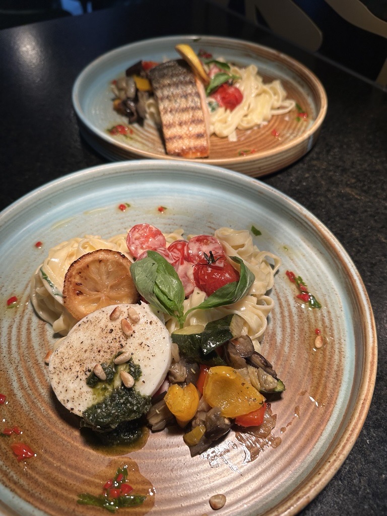 Two plates of pasta with grilled vegetables, cherry tomatoes, basil, a slice of lemon, and a round piece of mozzarella topped with pesto and pine nuts.