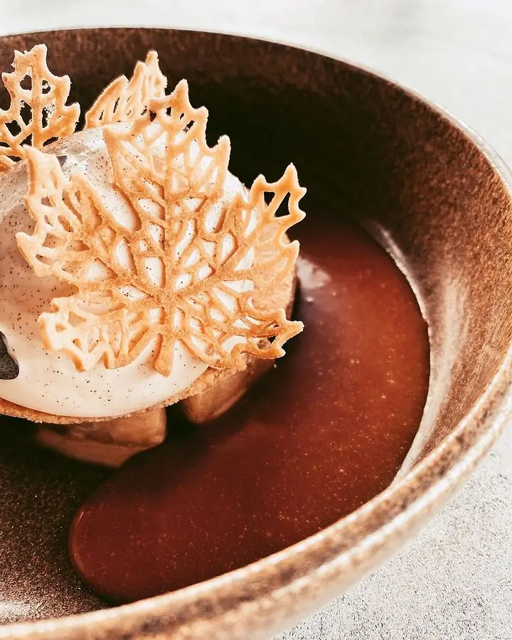 Elegant dessert with a scoop of cream topped with delicate leaf-shaped cookie decorations, served with a pool of chocolate sauce in a brown bowl.