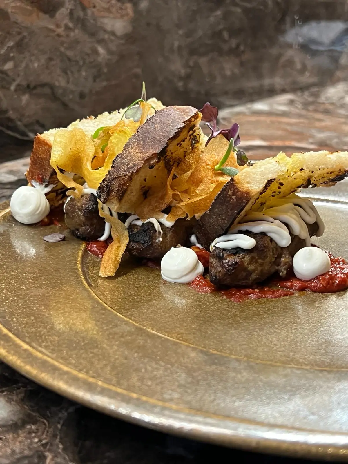 Gourmet dish with grilled meat topped with toasted bread, crispy potato chips, white sauce, and microgreens on a ceramic plate.