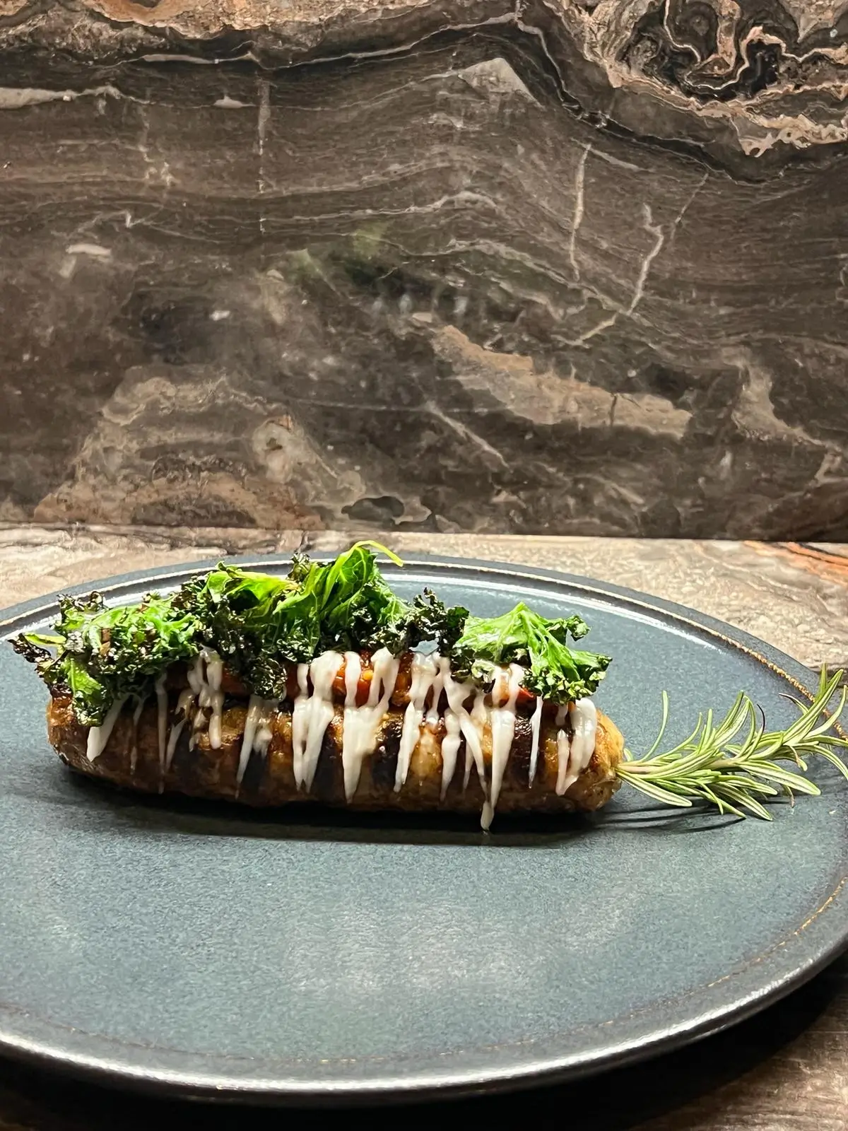 Stuffed baked potato topped with melted white sauce and crispy kale on a dark blue plate with a sprig of rosemary.