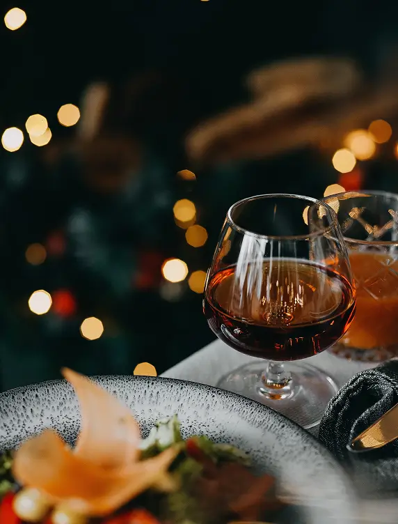 Close-up of a glass of amber-colored liquor on a table with another glass and a blurred plate of food, set against warm, out-of-focus lights.