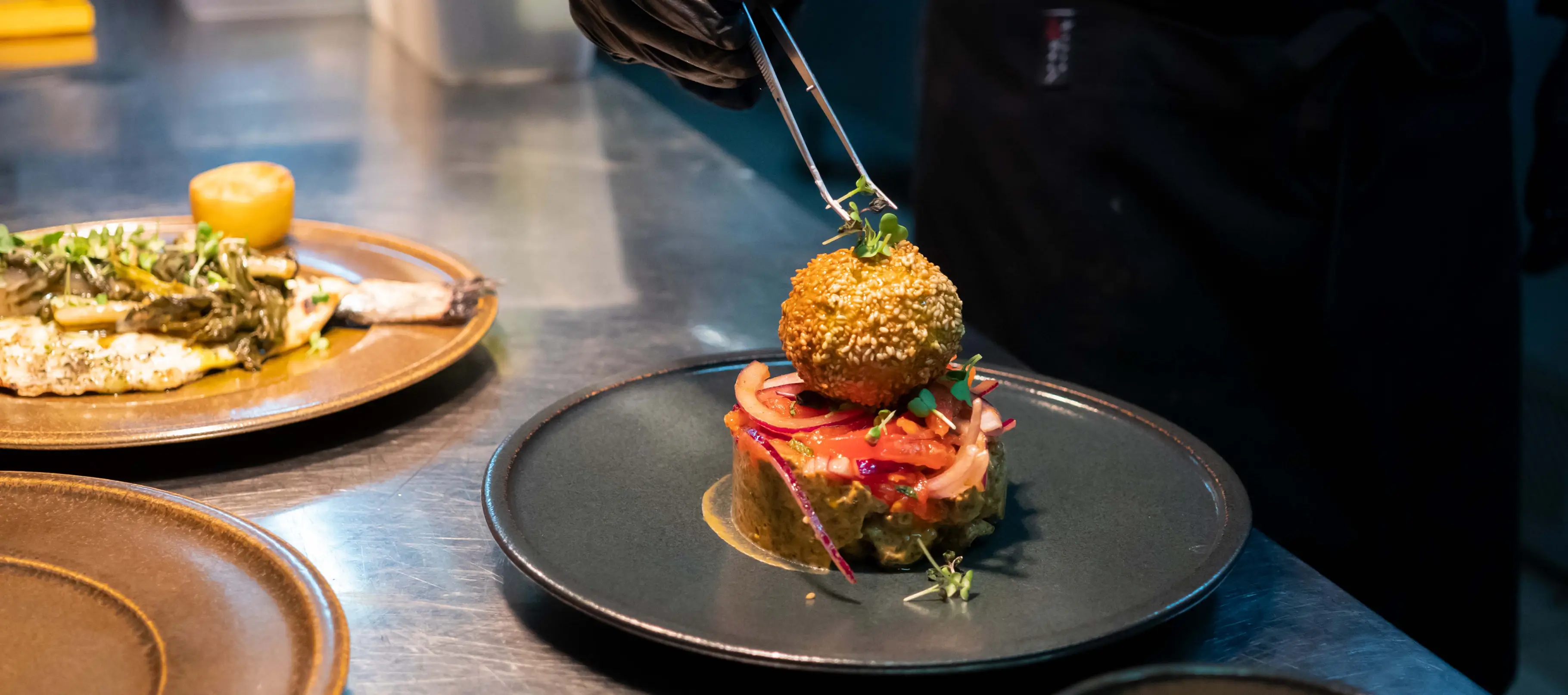 A chef using tongs to place microgreens on a sesame-crusted fried ball atop a dish garnished with sliced onions and microgreens on a black plate.