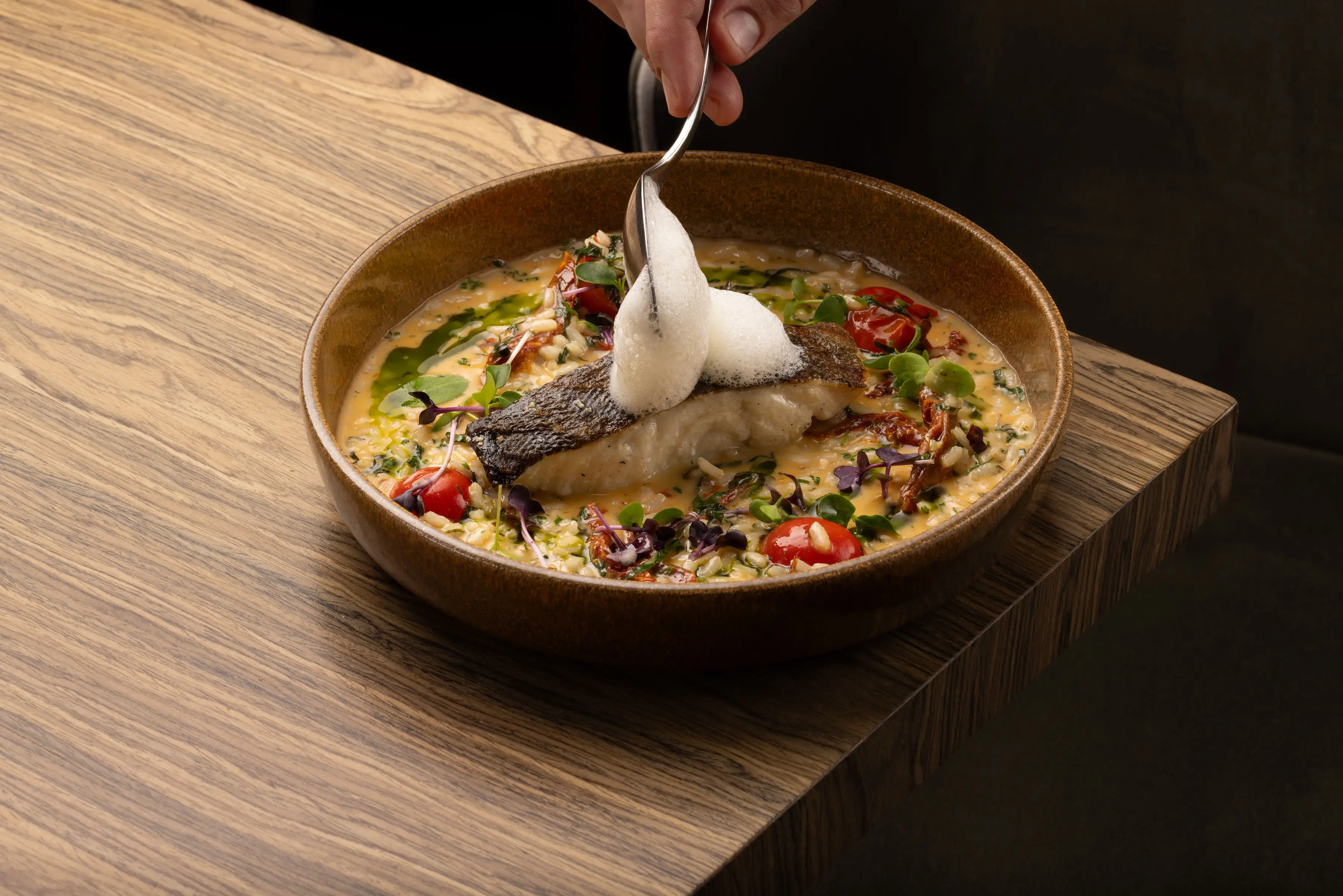 Seared fish fillet with foam on top served over a creamy risotto with cherry tomatoes and greens in a brown bowl on a wooden table.