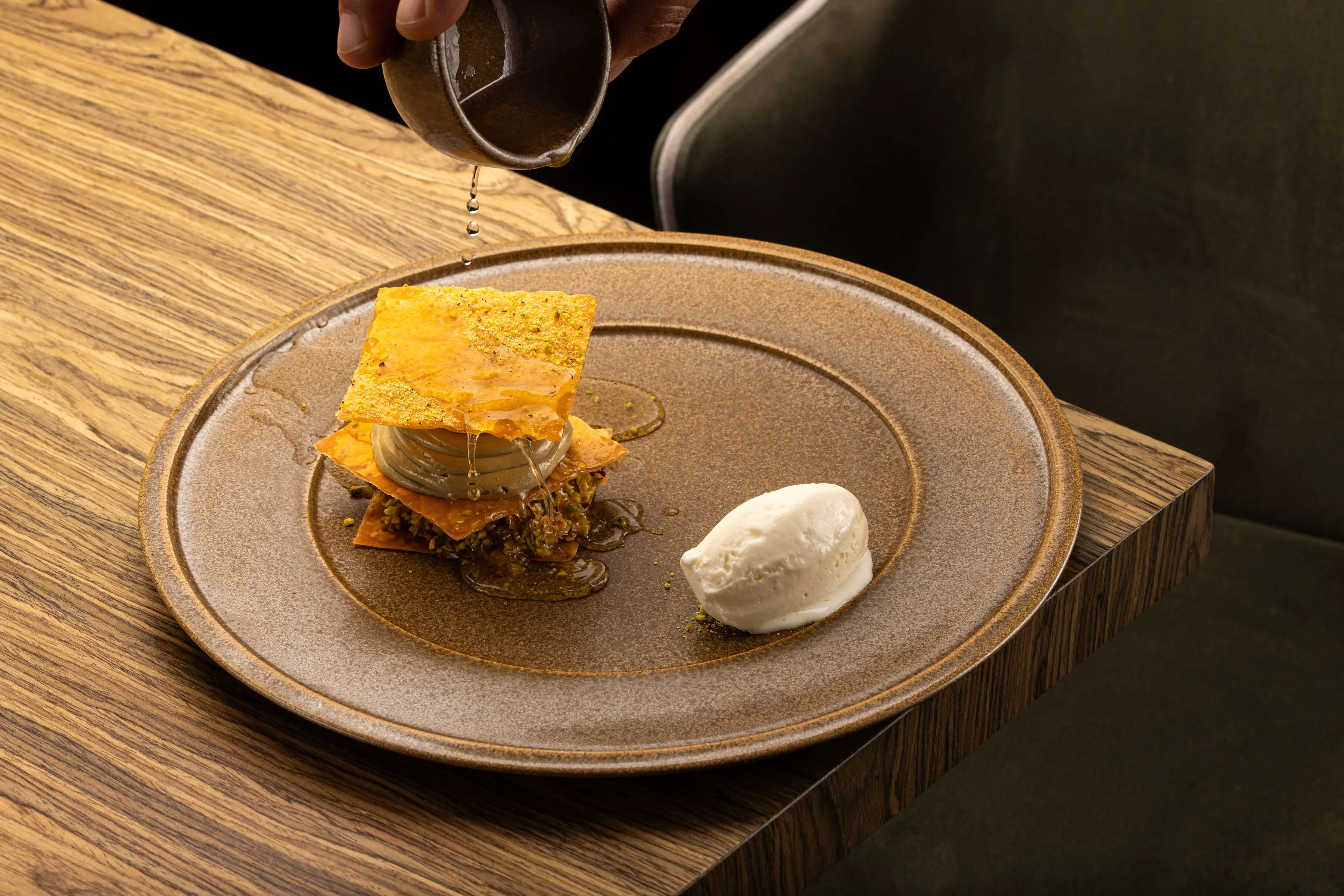Hand pouring syrup over a modern layered baklava dessert with ice cream on a brown plate.
