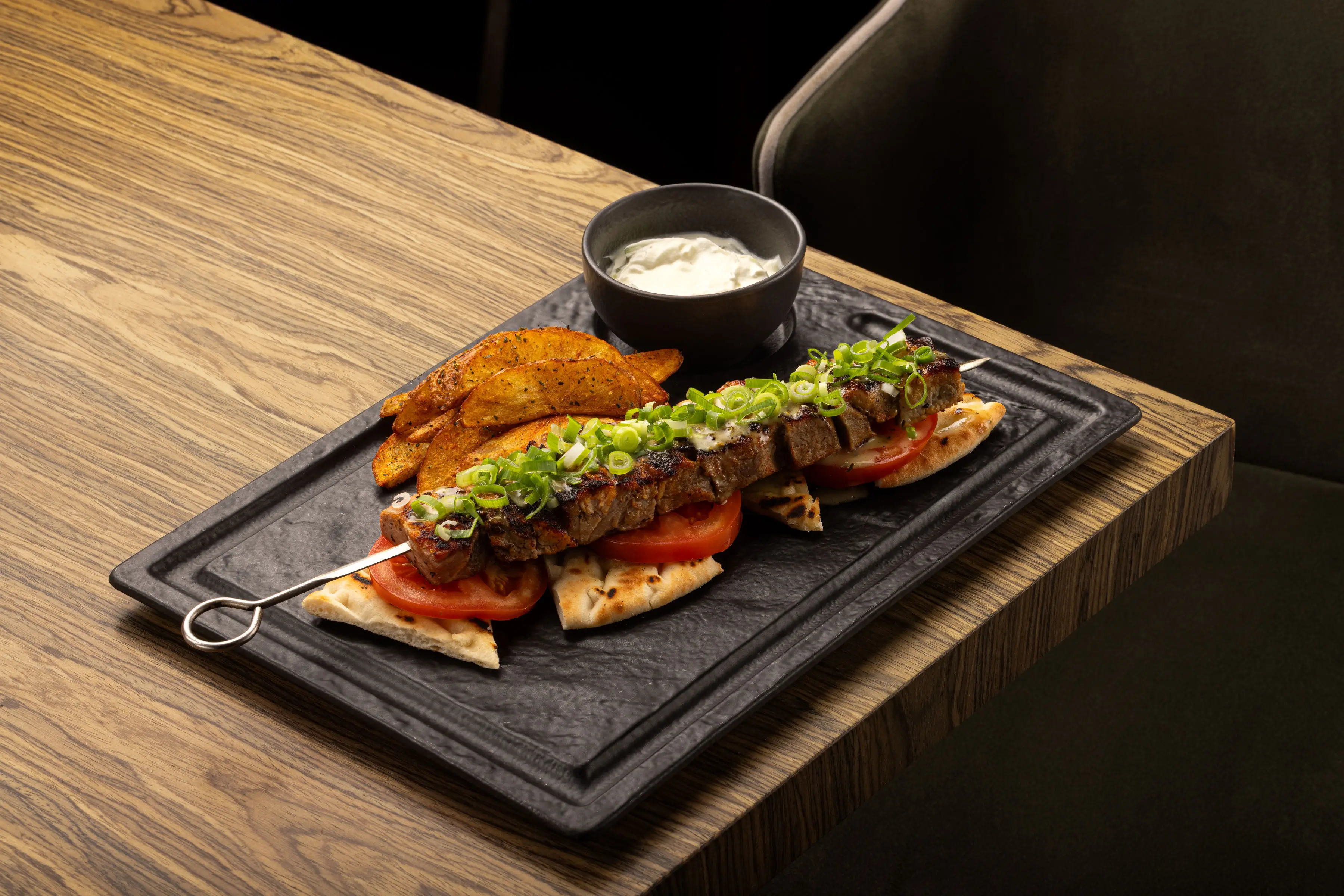 Greek souvlaki skewer with grilled meat topped with chopped green onions, served on pita bread with tomato slices, crispy potato wedges, and a bowl of white dipping sauce on a black rectangular plate.