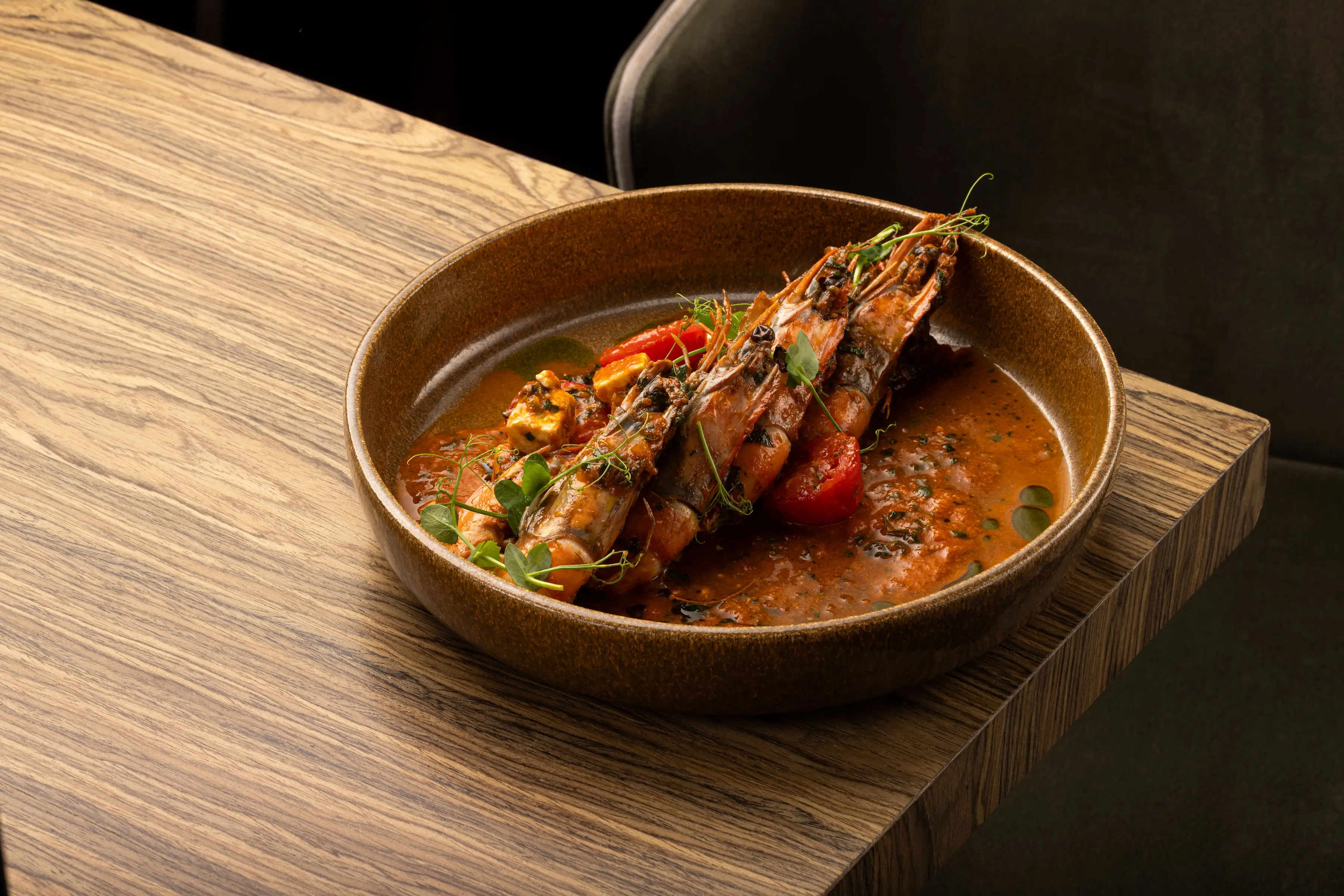 Brown bowl with three large cooked prawns in tomato-based sauce, garnished with herbs and vegetables on a wooden table.