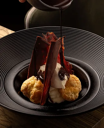 Elegant dessert with cream, cookies, and dark chocolate shards on a black plate being drizzled with chocolate sauce.