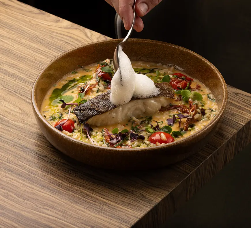 Hand spooning foam onto a plated dish of seared fish over creamy risotto with cherry tomatoes and greens in a brown bowl on a wooden table.