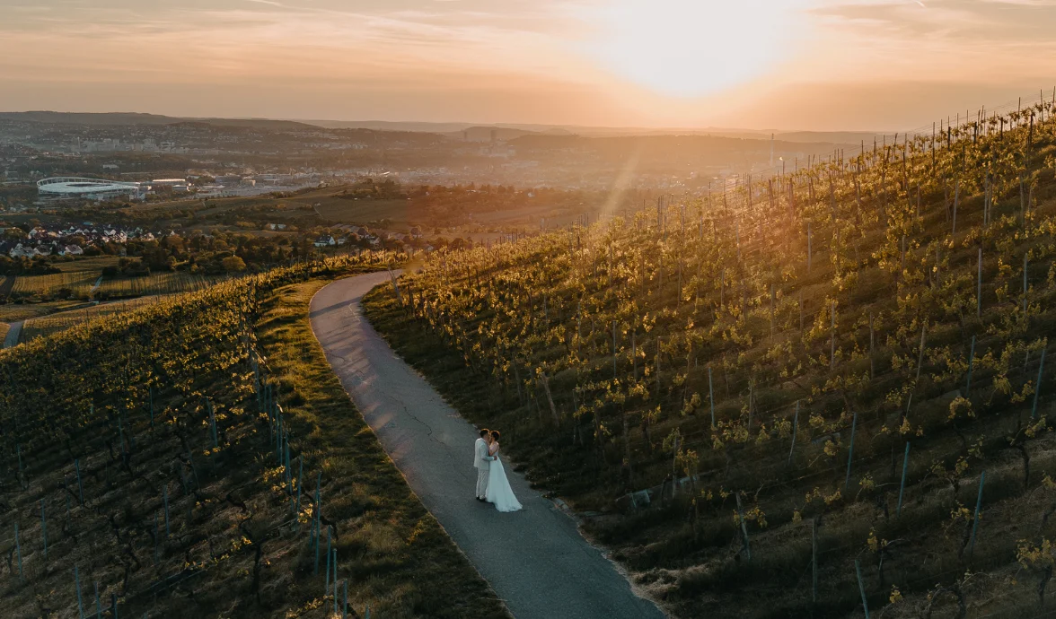 Bride and groom embracing on a winding vineyard road at sunset with a cityscape in the background.