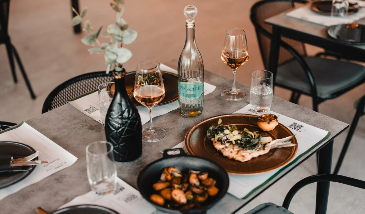 Restaurant table with a grilled fish dish, a bowl of roasted vegetables, two glasses of rosé wine, a bottle of water, and a black vase with green leaves.