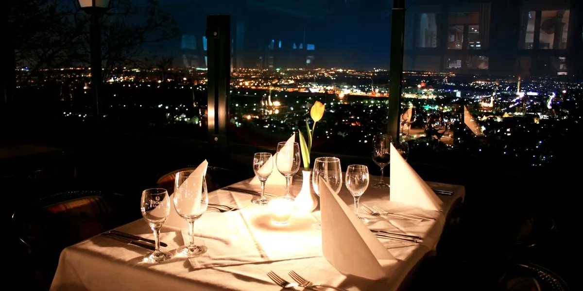 Elegant dining table set with white napkins, wine glasses, and a single yellow tulip vase overlooking a city skyline at night.