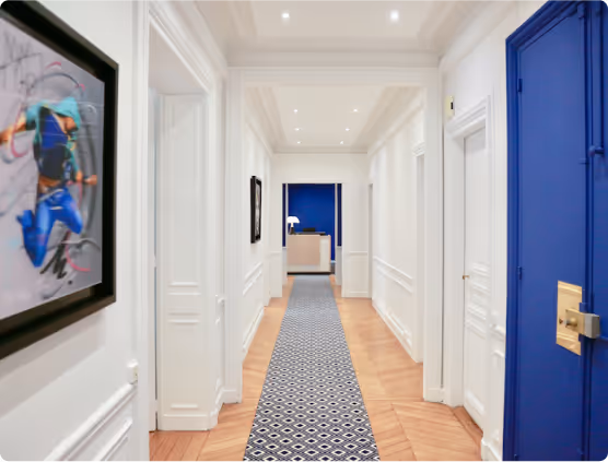 Couloir long et éclairé avec murs blancs, sol en bois clair, tapis bleu à motifs géométriques et porte bleue à droite.