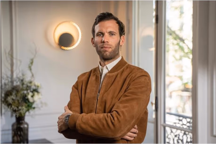 Homme aux cheveux courts portant une veste marron, debout bras croisés dans une pièce lumineuse avec une fenêtre et une plante en arrière-plan.