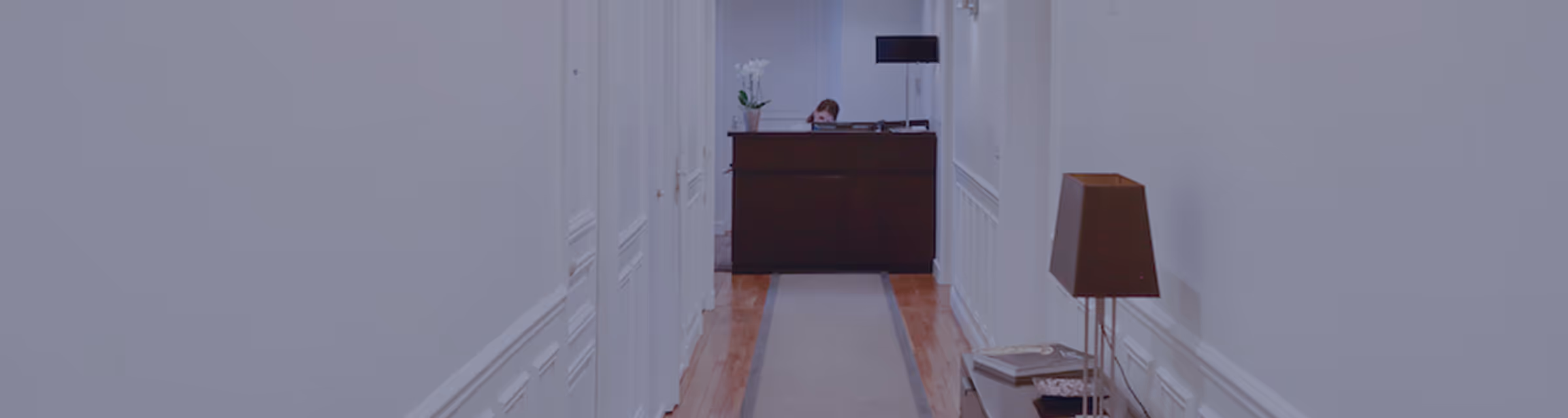 Couloir blanc avec un tapis gris menant à un bureau sombre où une personne est assise derrière la réception.