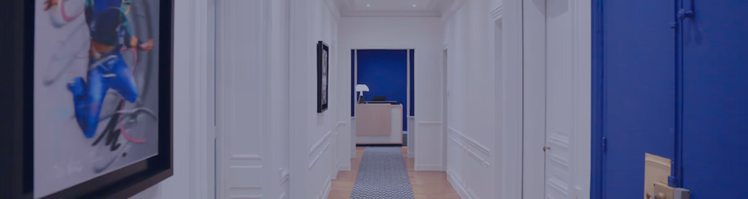 Couloir blanc éclairé avec tapis gris menant à un comptoir d'accueil devant un mur bleu.