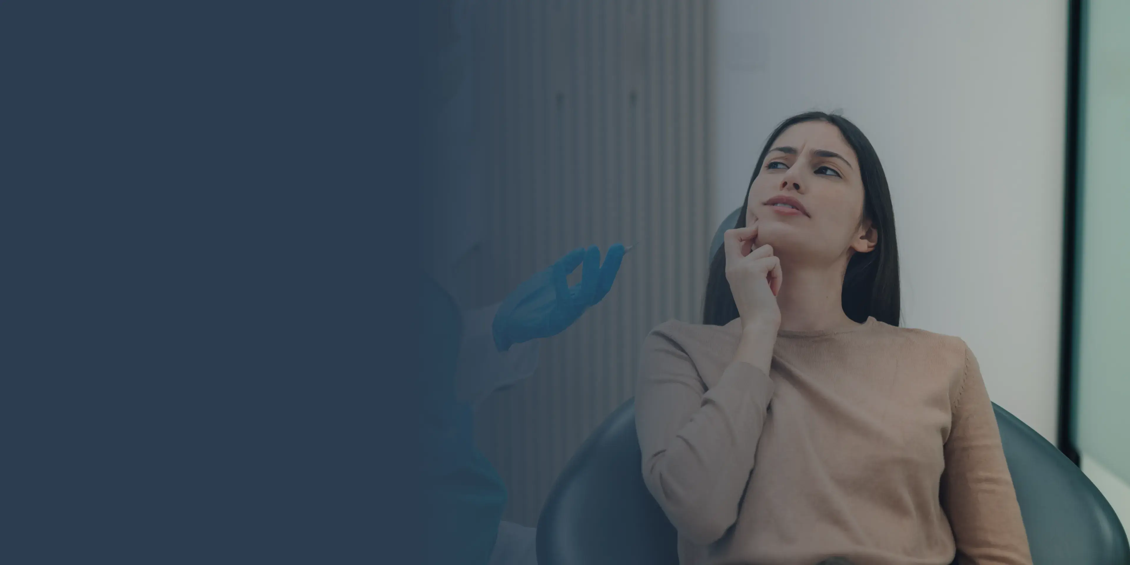 Woman sitting in dentist chair, touching cheek with a concerned expression.