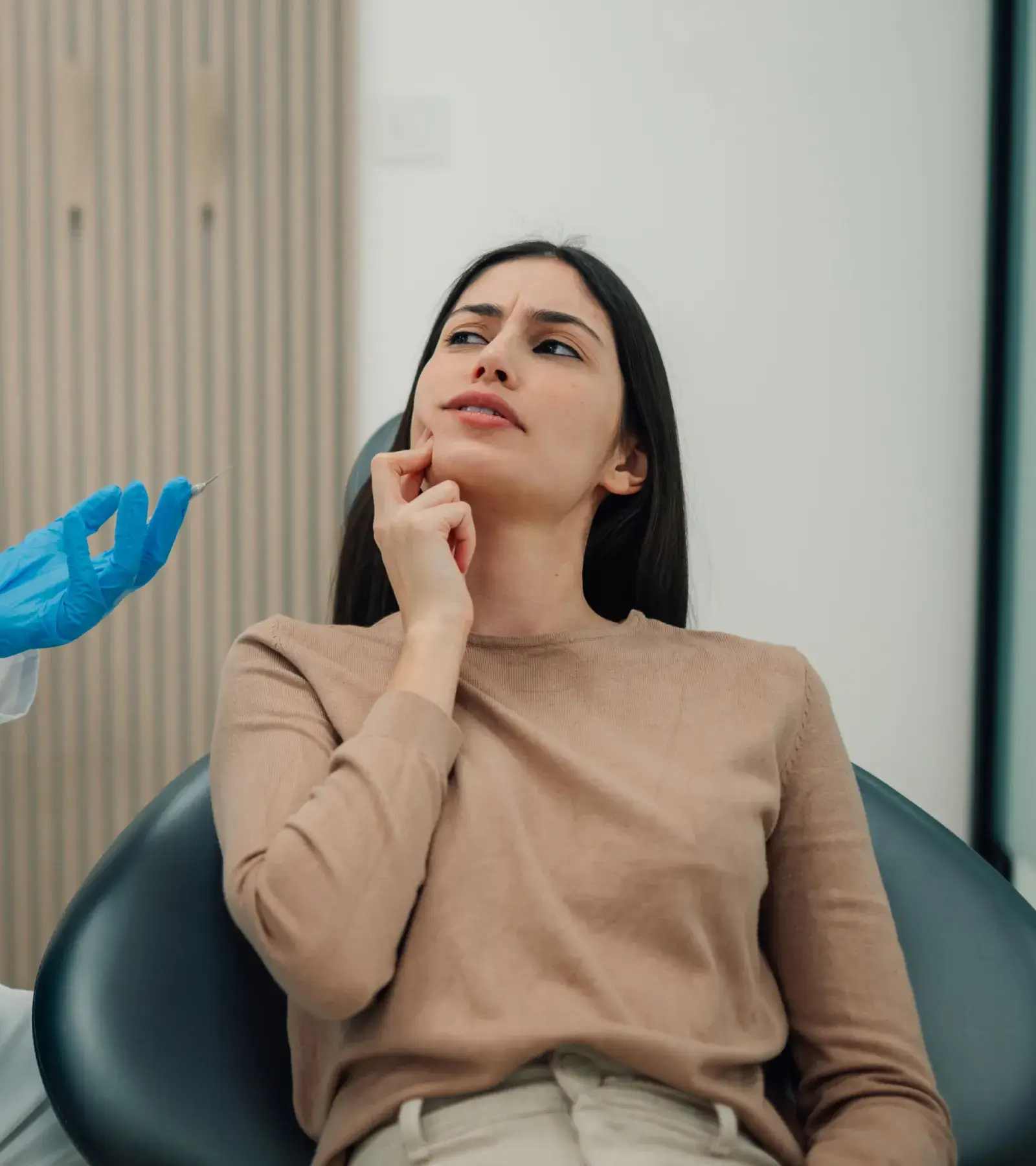 Woman sitting in a chair touching her jaw, looking at a gloved hand holding a syringe.