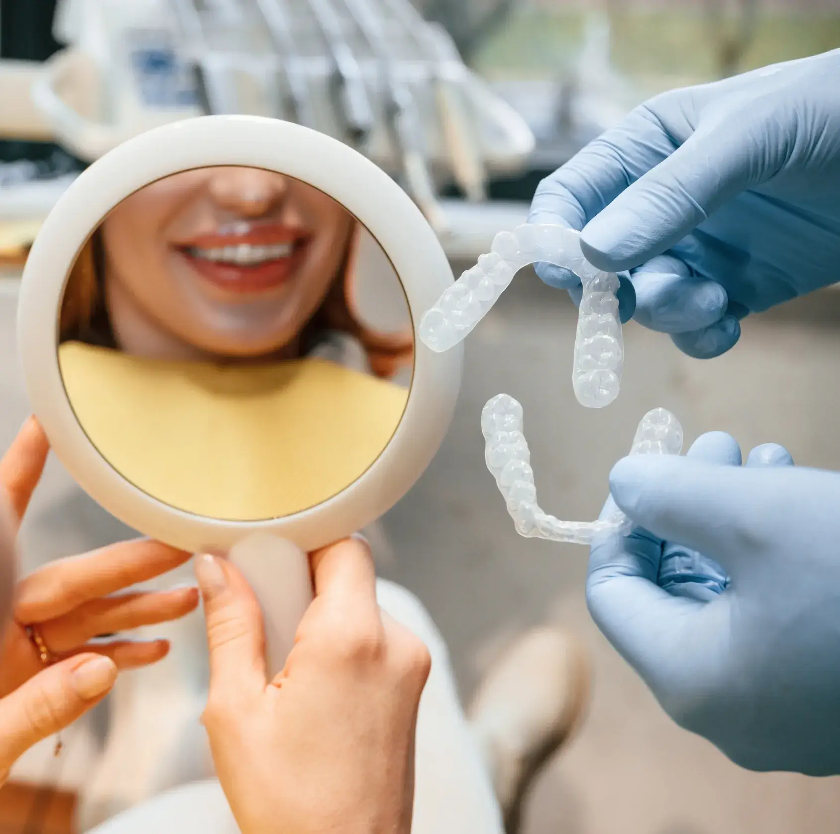 Woman holding mirror smiling while dentist shows clear dental aligners with gloved hands.