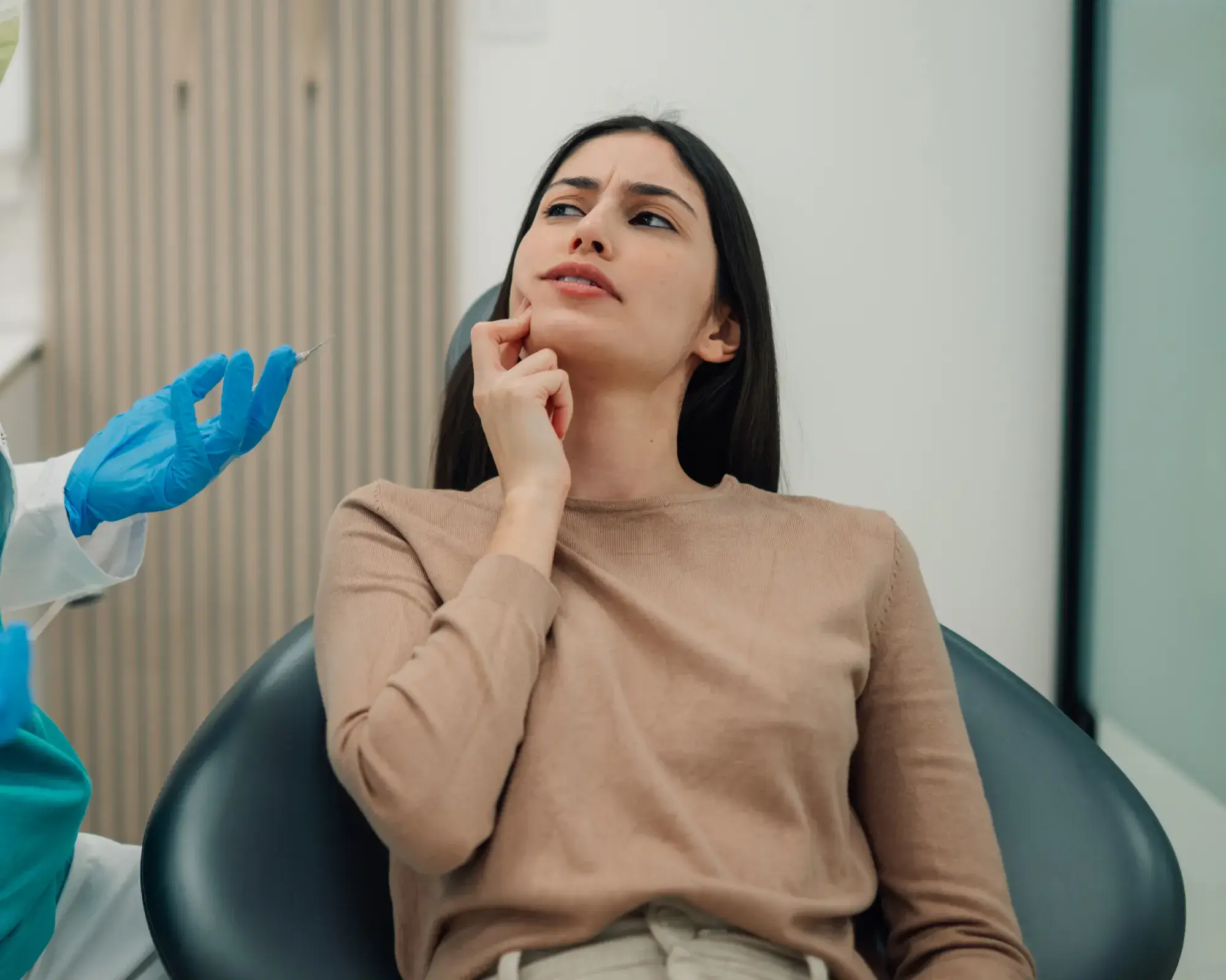 Woman in beige shirt touching her jaw with worried face, person in blue gloves holding dental syringe.