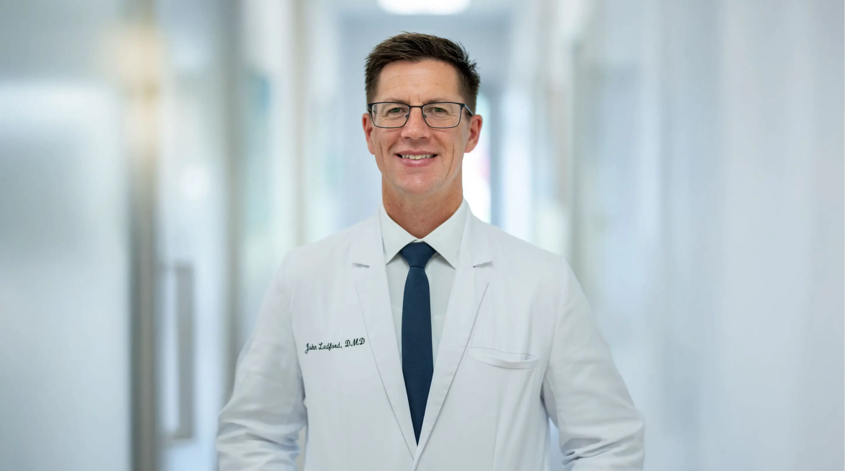 Smiling man wearing glasses and a white coat with a navy tie standing in a bright hallway.