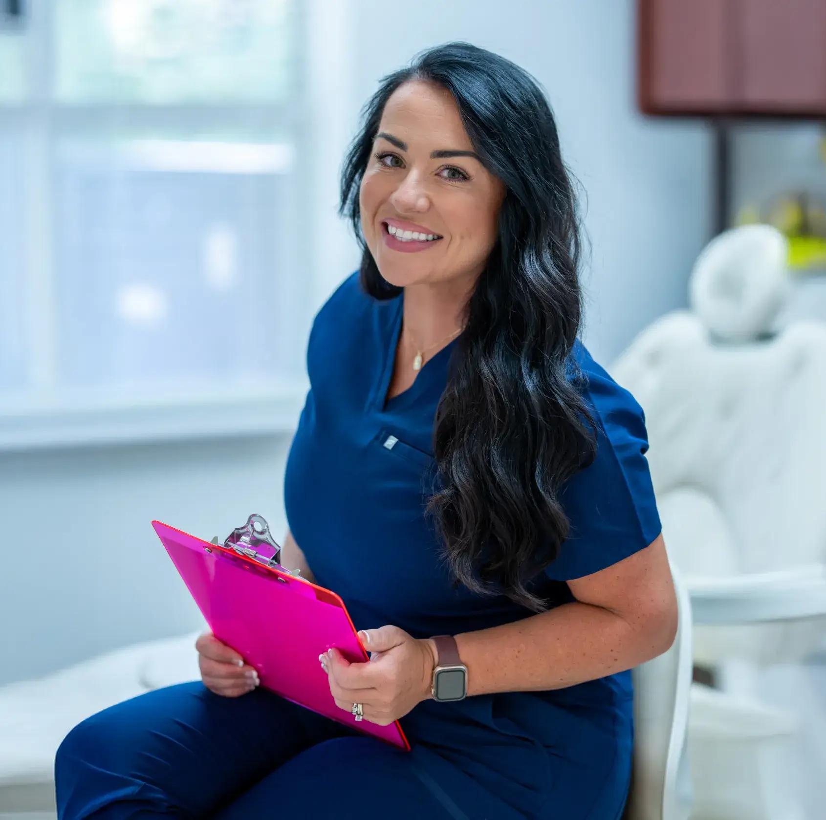 Smiling female healthcare worker in blue scrubs holding a pink clipboard in a bright room.