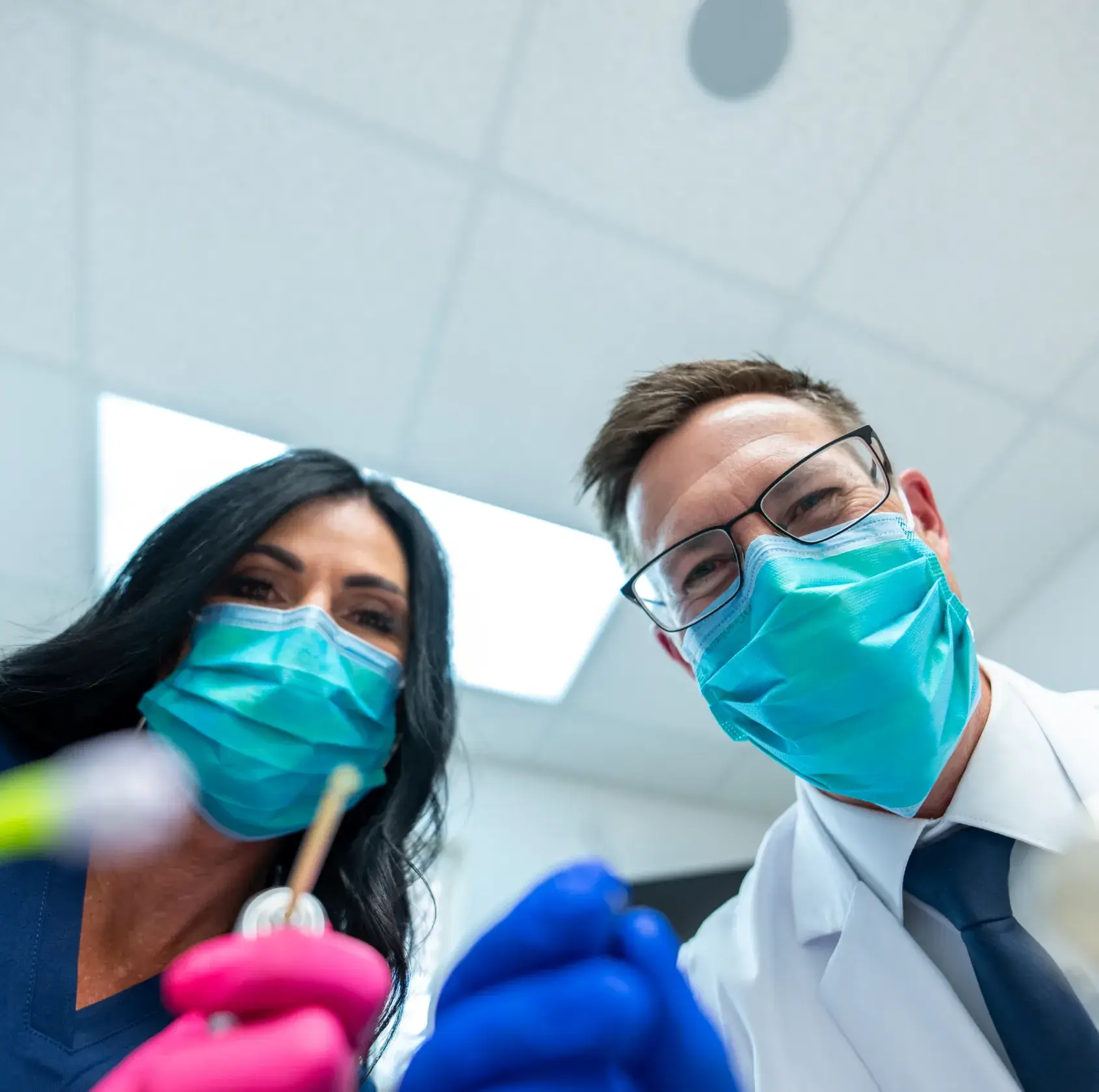 Two masked dentists, one in gloves, holding dental tools, looking down at the camera.