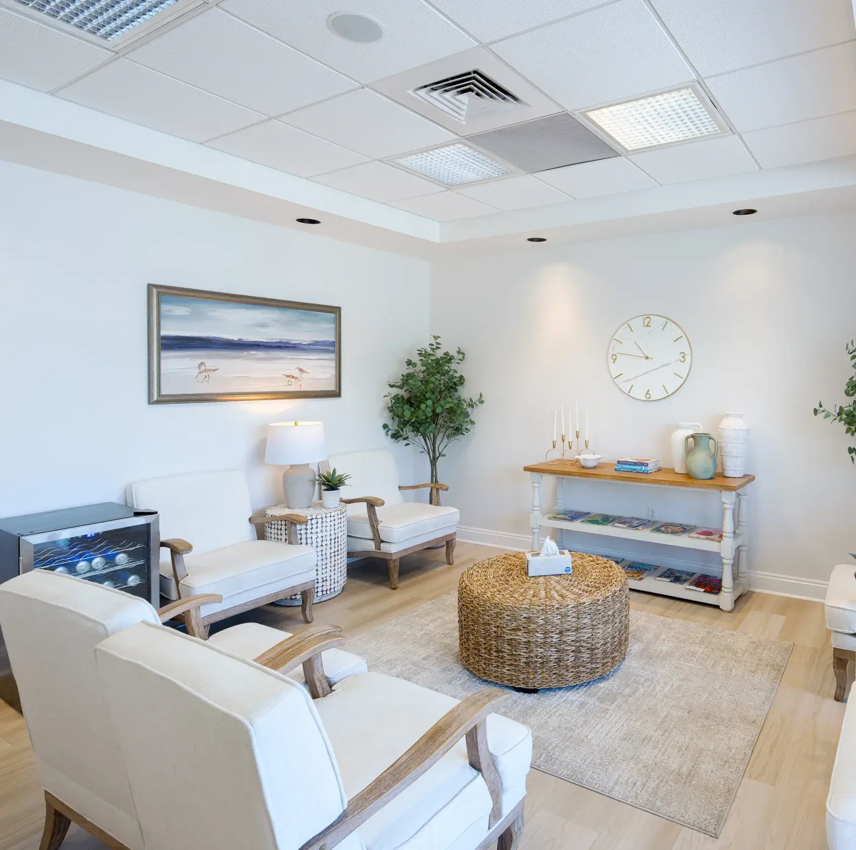 Bright waiting room with cream chairs, woven coffee table, bookshelves, and coastal artwork on the wall.