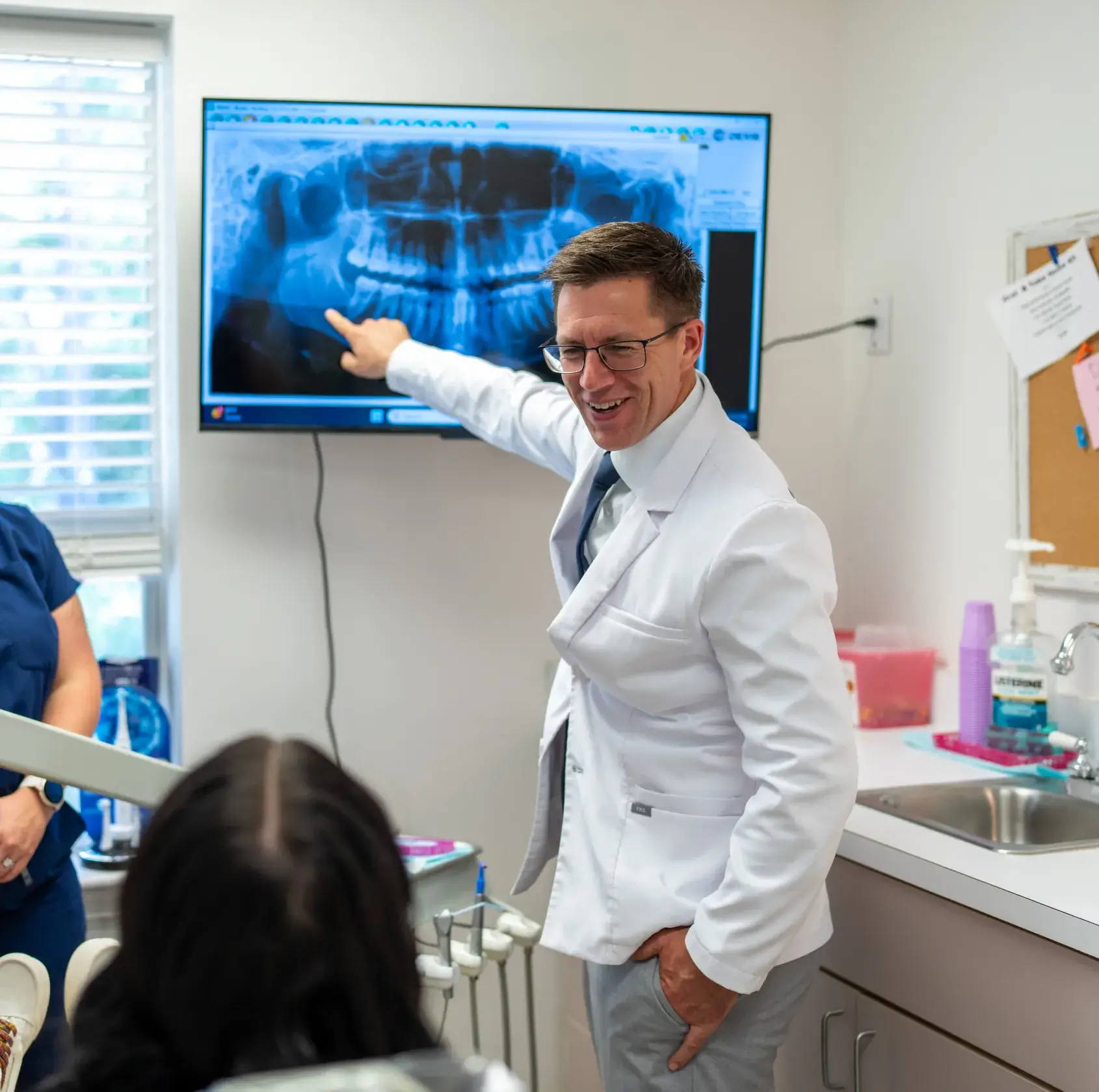 Dentist in white coat pointing to a dental X-ray displayed on a screen for a patient in the clinic.