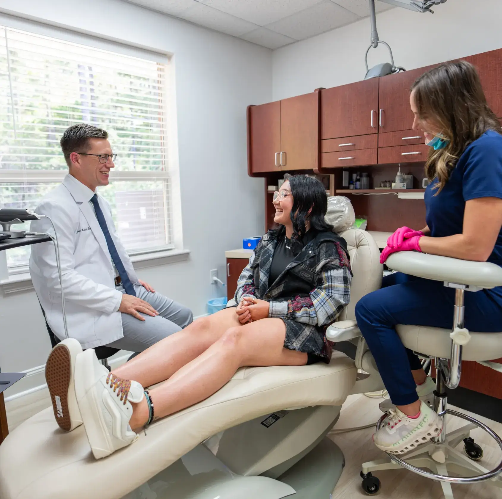 A female patient sits and smiles in a dental chair as a dentist and hygienist talk to her in the office.