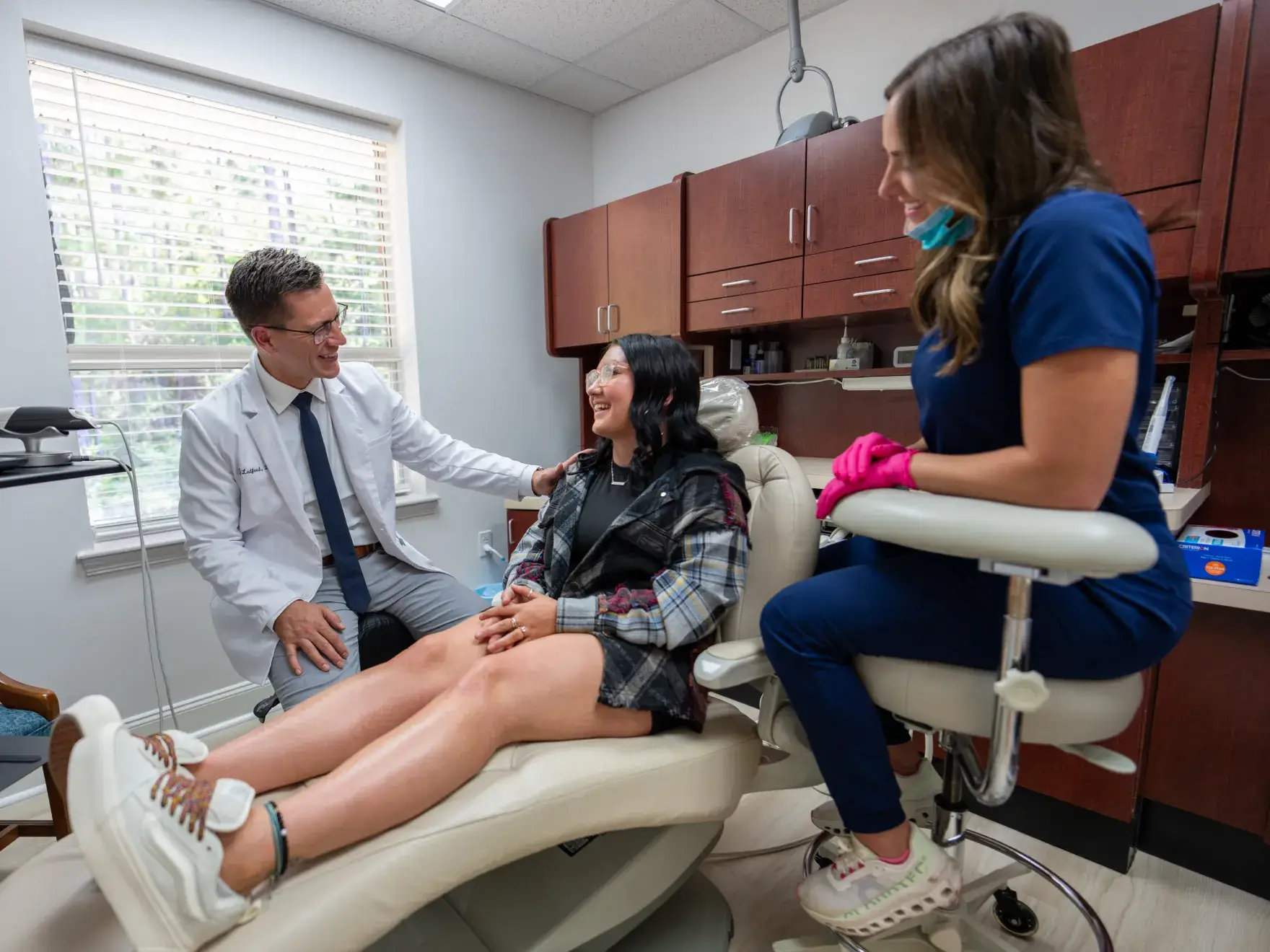 Dentist and dental assistant smiling and talking with a female patient seated in a dental chair.
