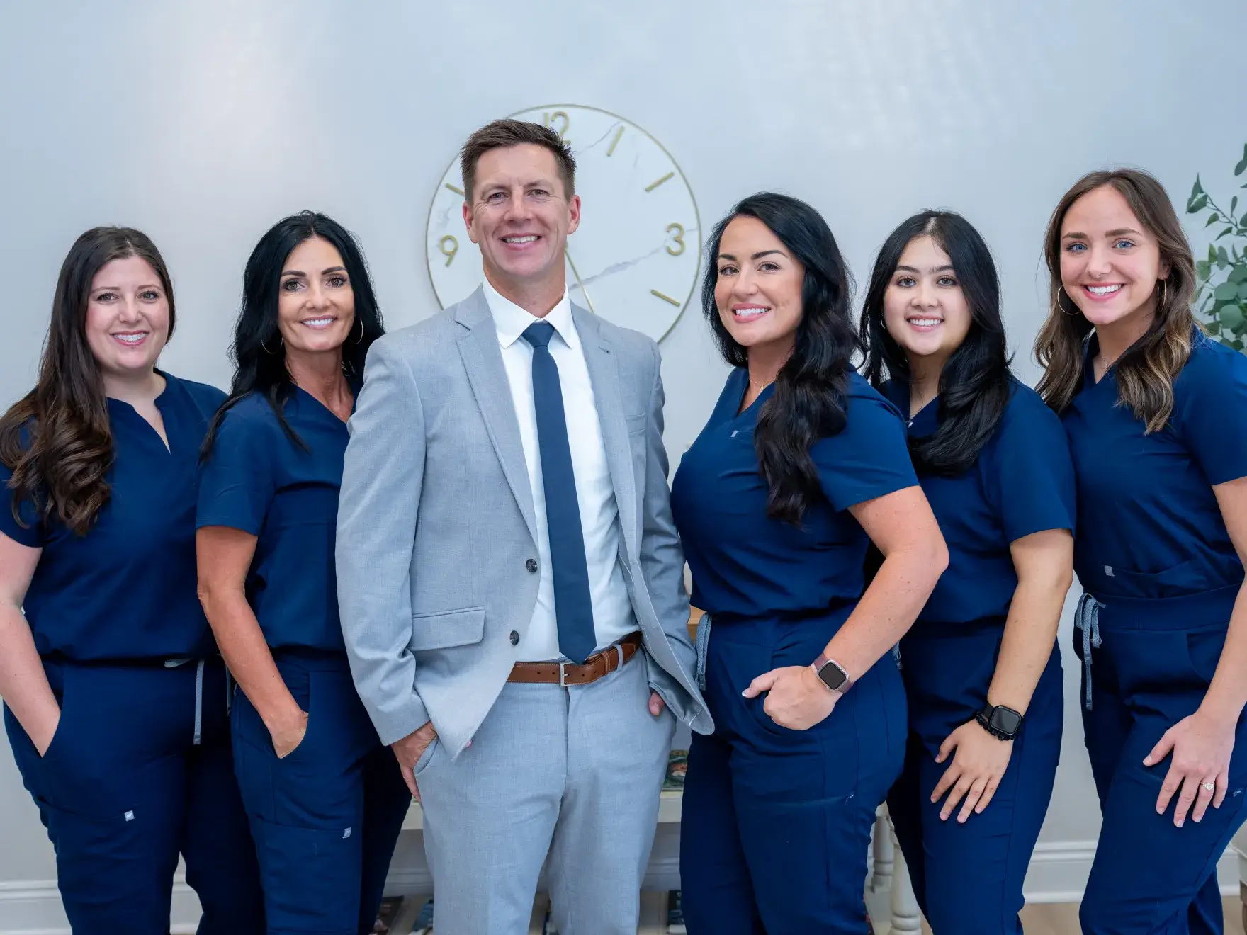 Six smiling dental professionals including five women in navy scrubs and one man in a gray suit with a tie.