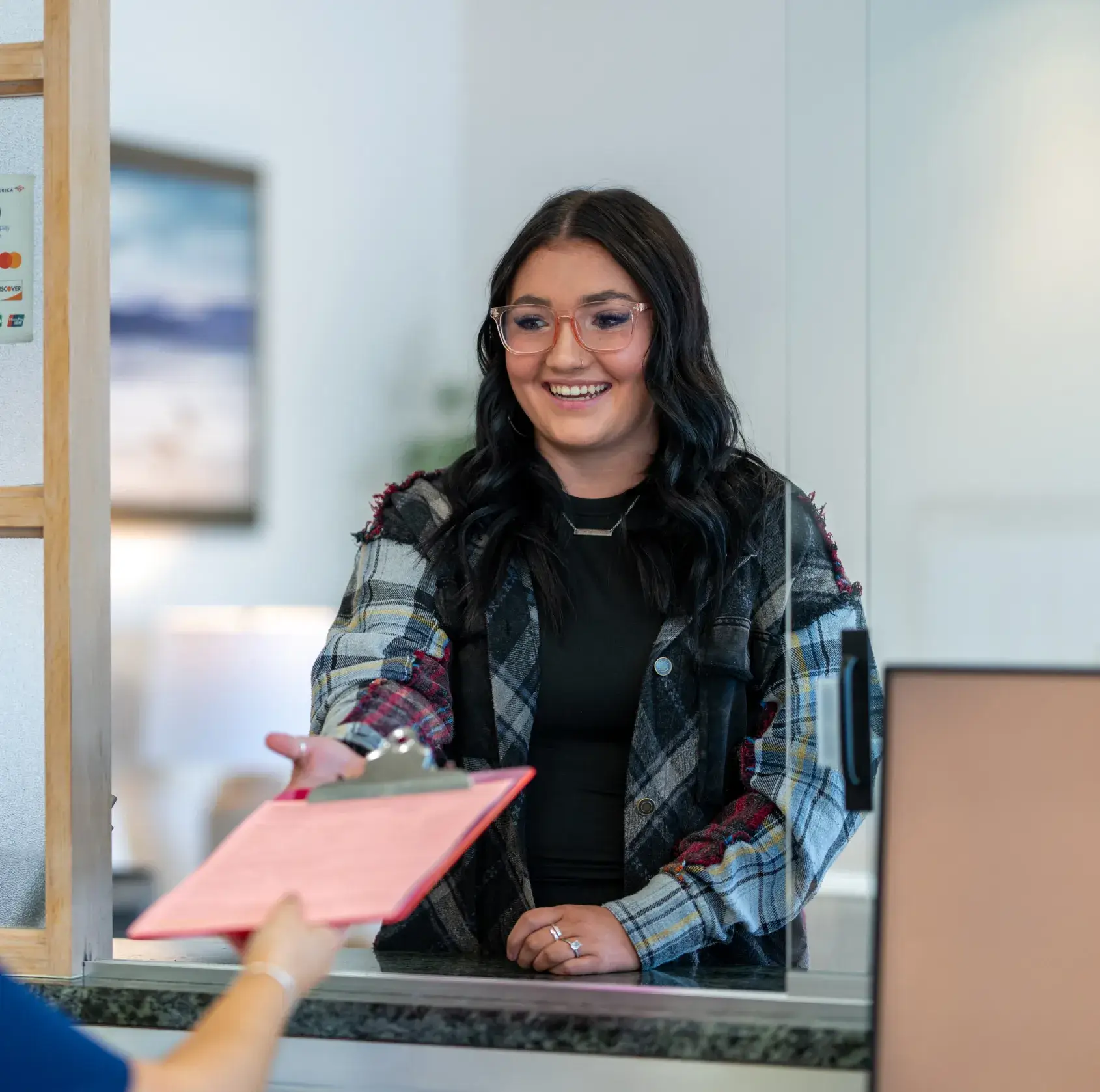 Smiling woman with glasses and plaid jacket receiving a clipboard over a counter window.