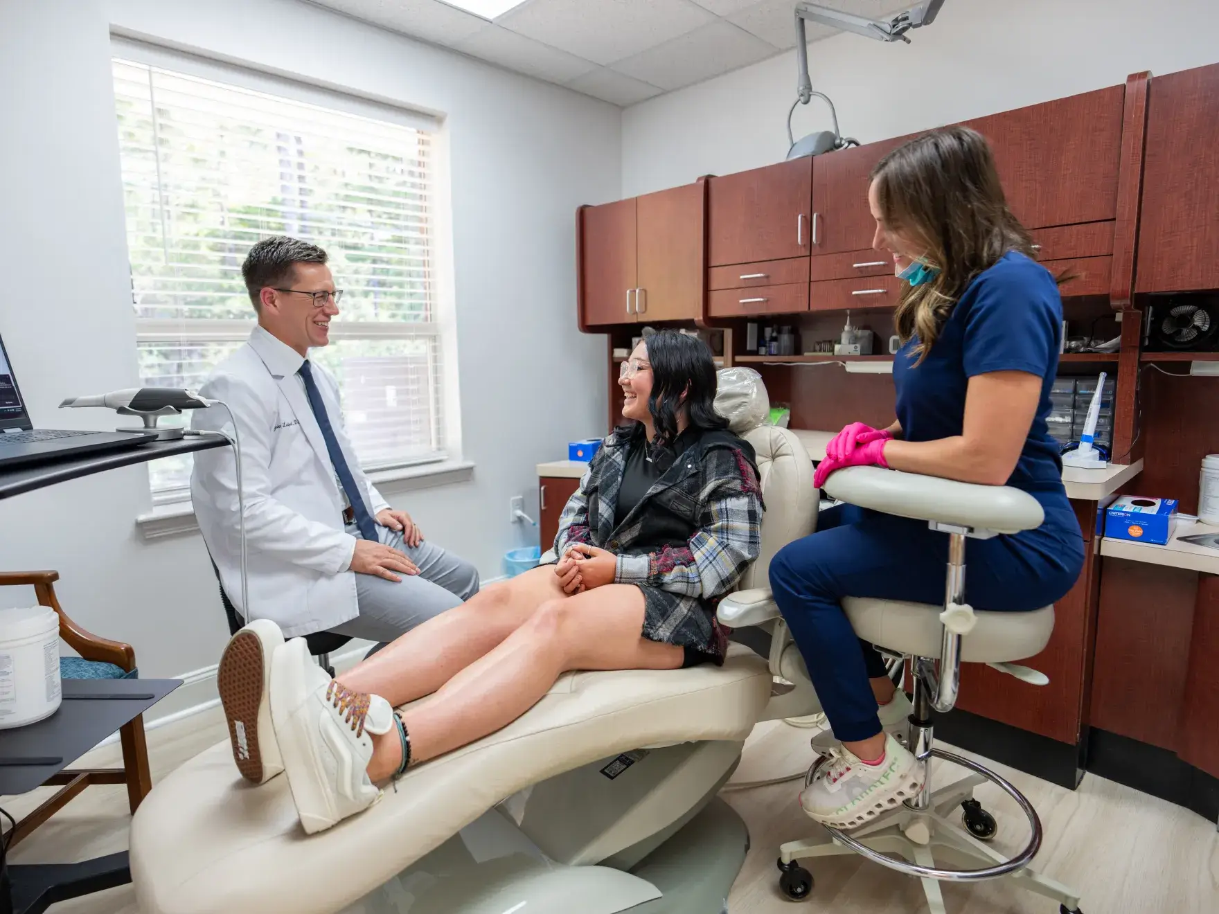 Woman patient in dental chair talking with male dentist and female assistant in clinic room.