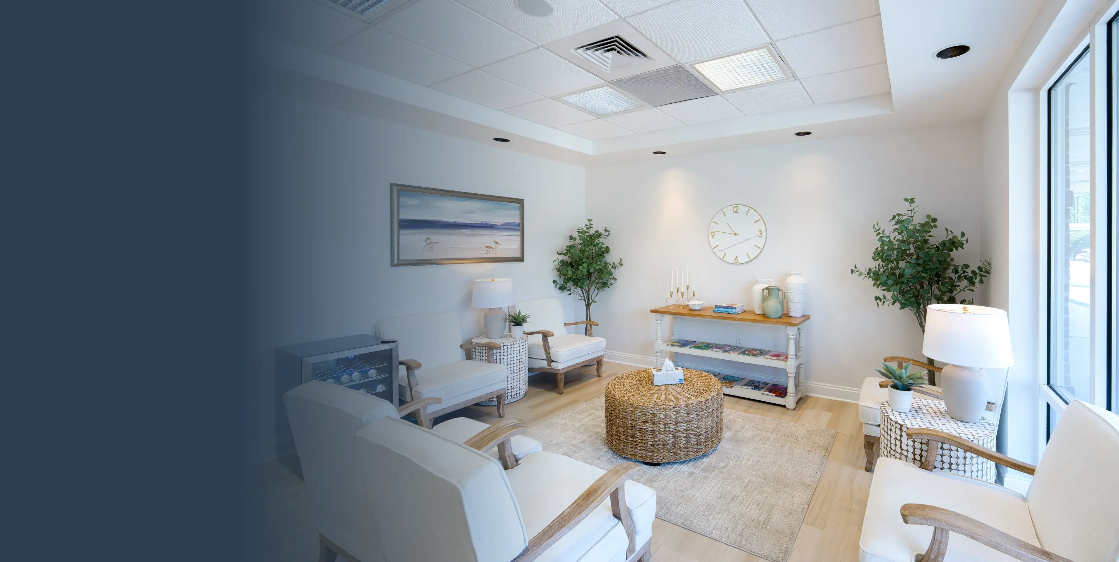 Bright waiting room with white chairs, wicker table, plants, wall clock, and large window.