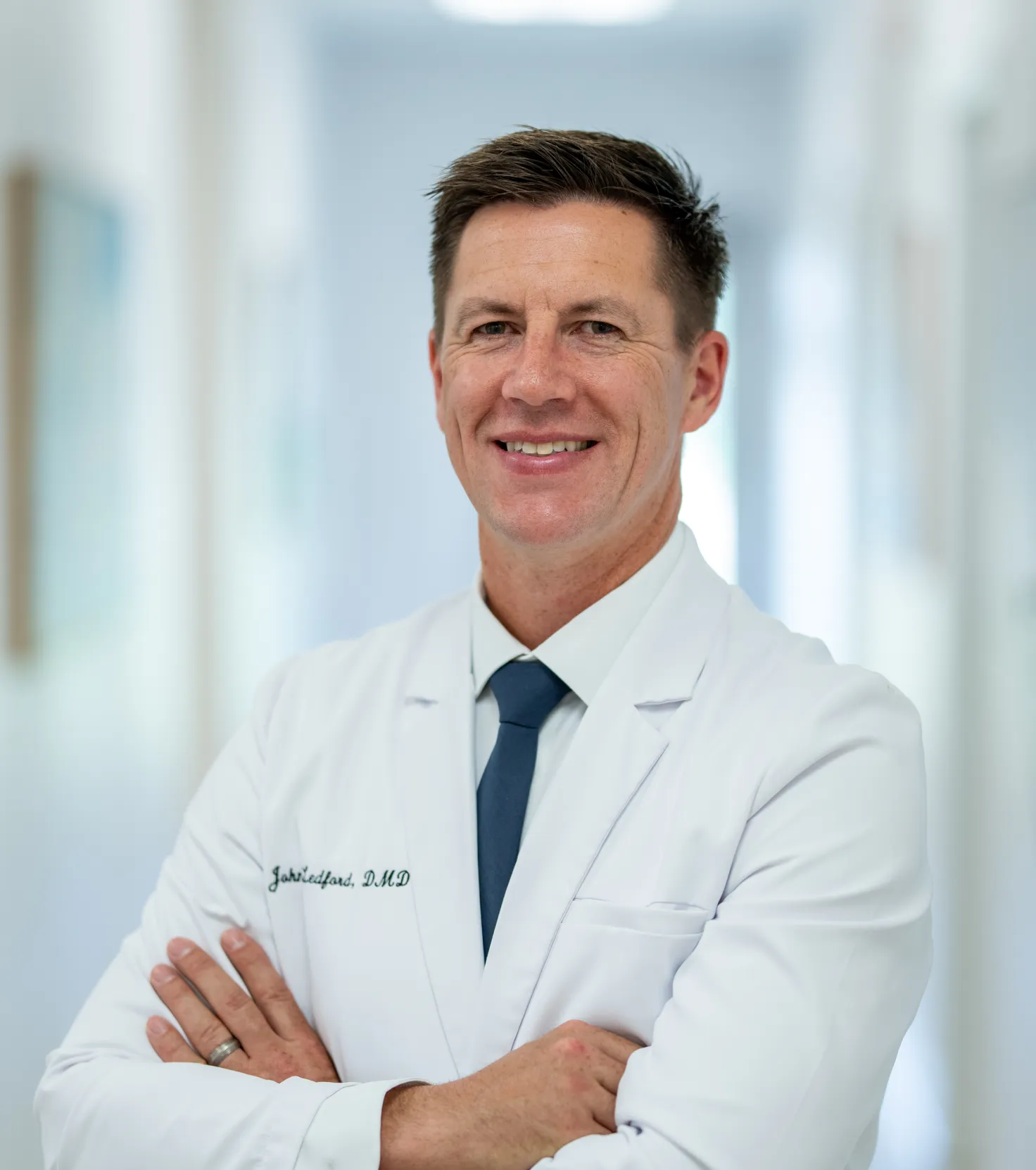 Smiling male doctor in white coat and tie with arms crossed standing in a bright hallway.