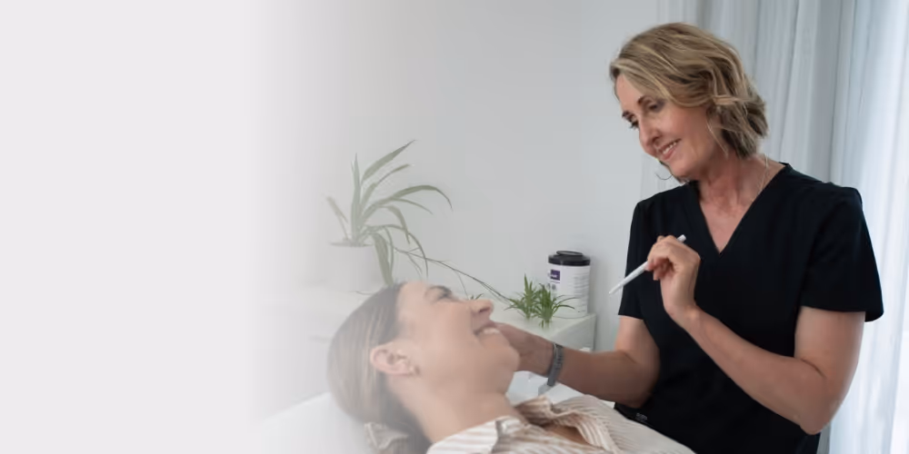 Smiling Christchurch appearance medicine professional in black scrubs holding a pen and interacting with a relaxed patient lying down.